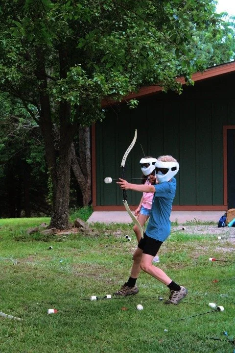 Two children wearing white masks in a backyard, one is holding a bow and aiming an arrow, surrounded by scattered golf balls, trees, and a shed in the background.