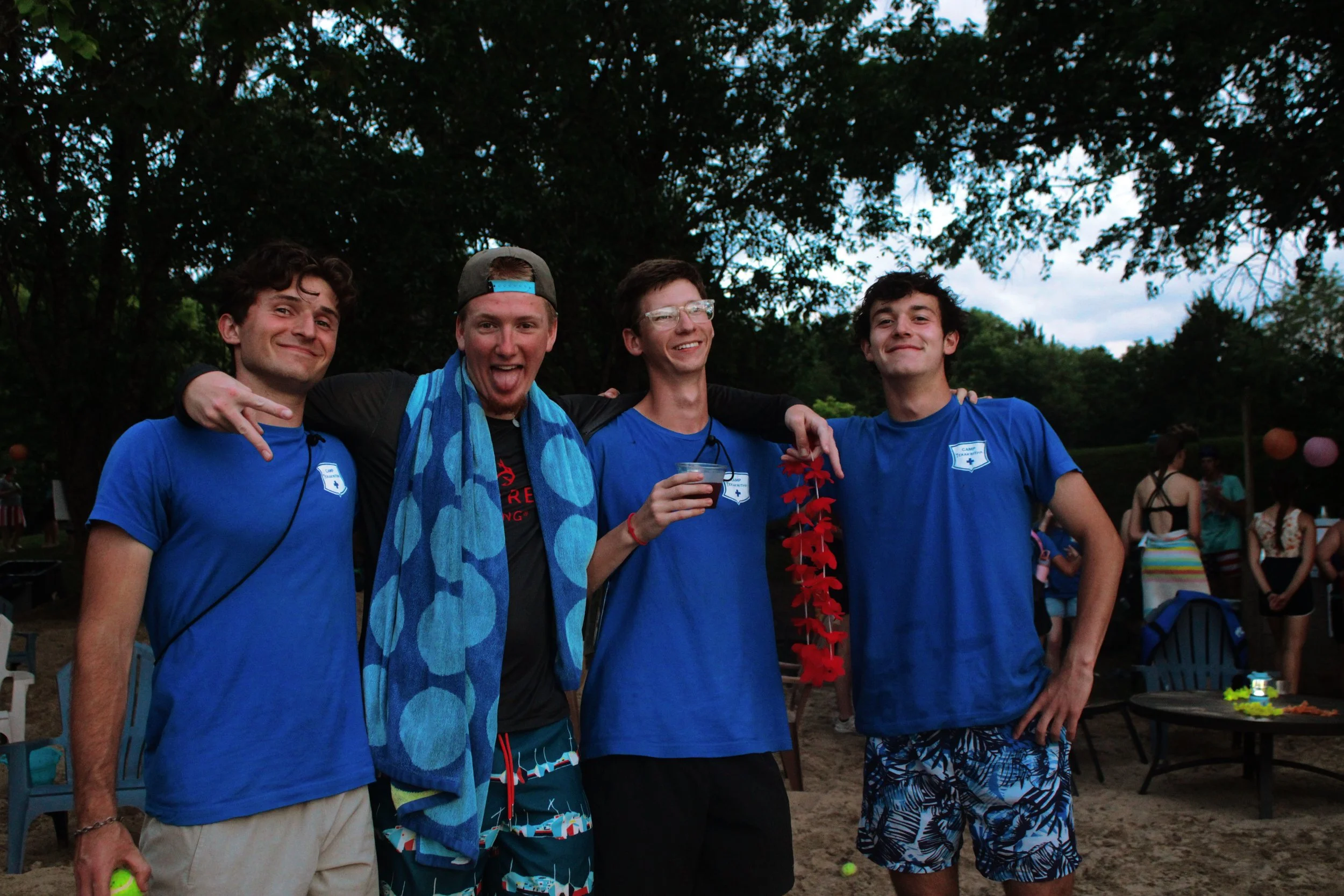 Four young men standing outdoors, smiling and posing together. They are wearing blue shirts, with a person in the middle holding a drink. The background includes trees and other people, suggesting a social or recreational event.