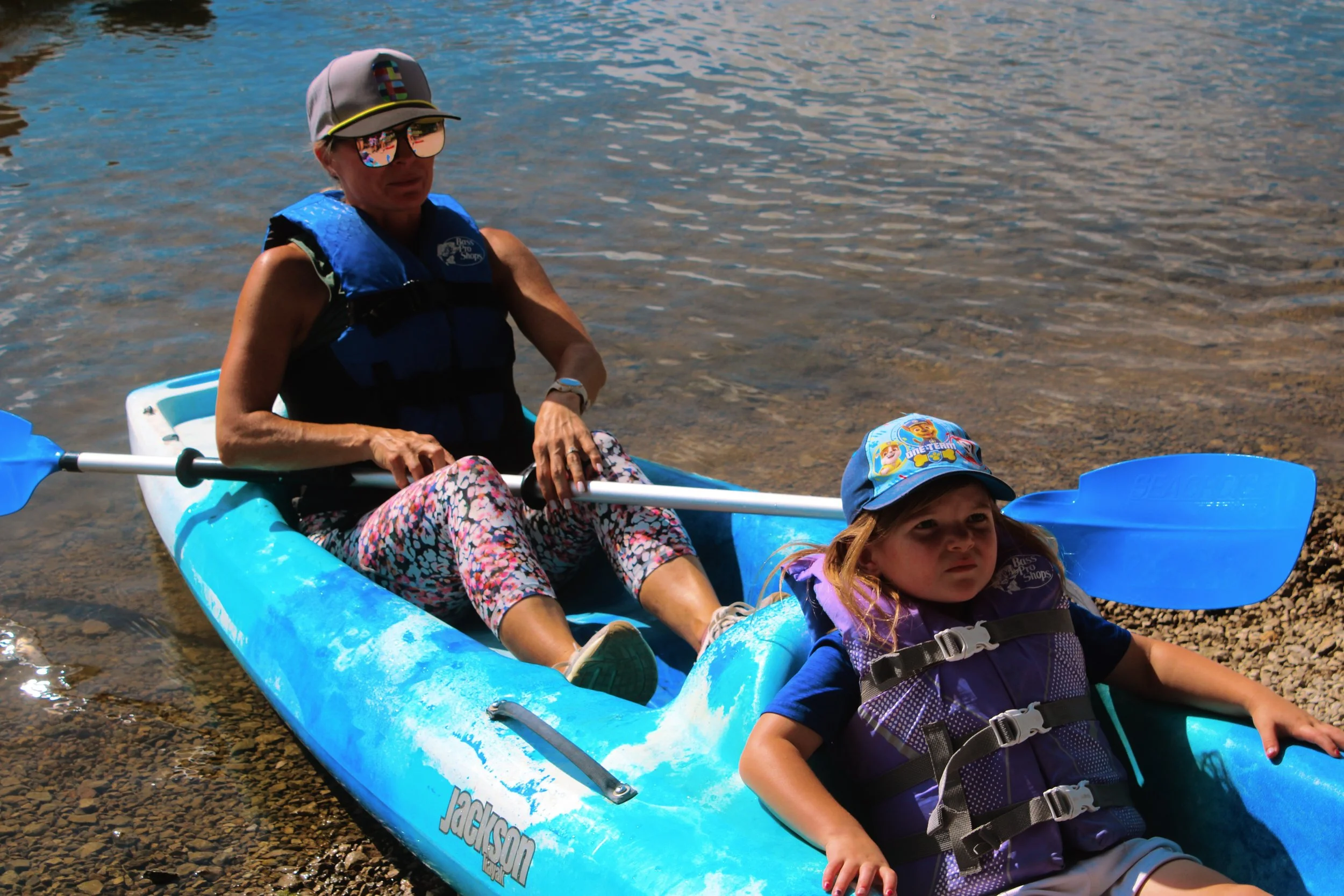 A woman and two children sitting in a blue kayak on shallow water near a pebbled shore.