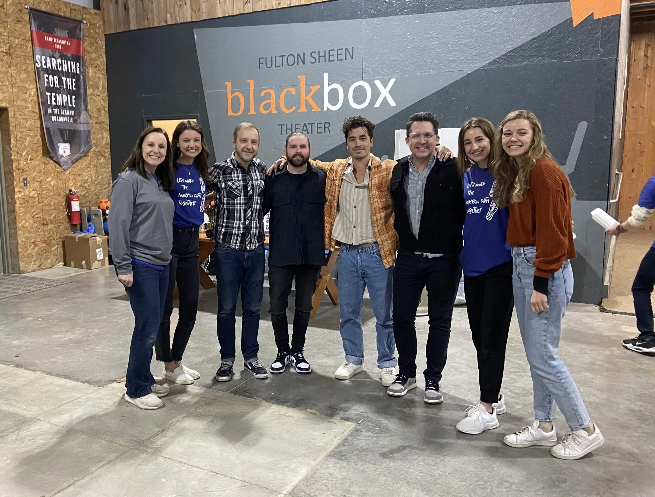 Seven people standing in a row in front of a large gray wall with the words "Fulton Sheen black box Theater" written on it. The group is smiling, and some are wearing matching blue shirts that say "Let's walk the narrow path together." The setting ap