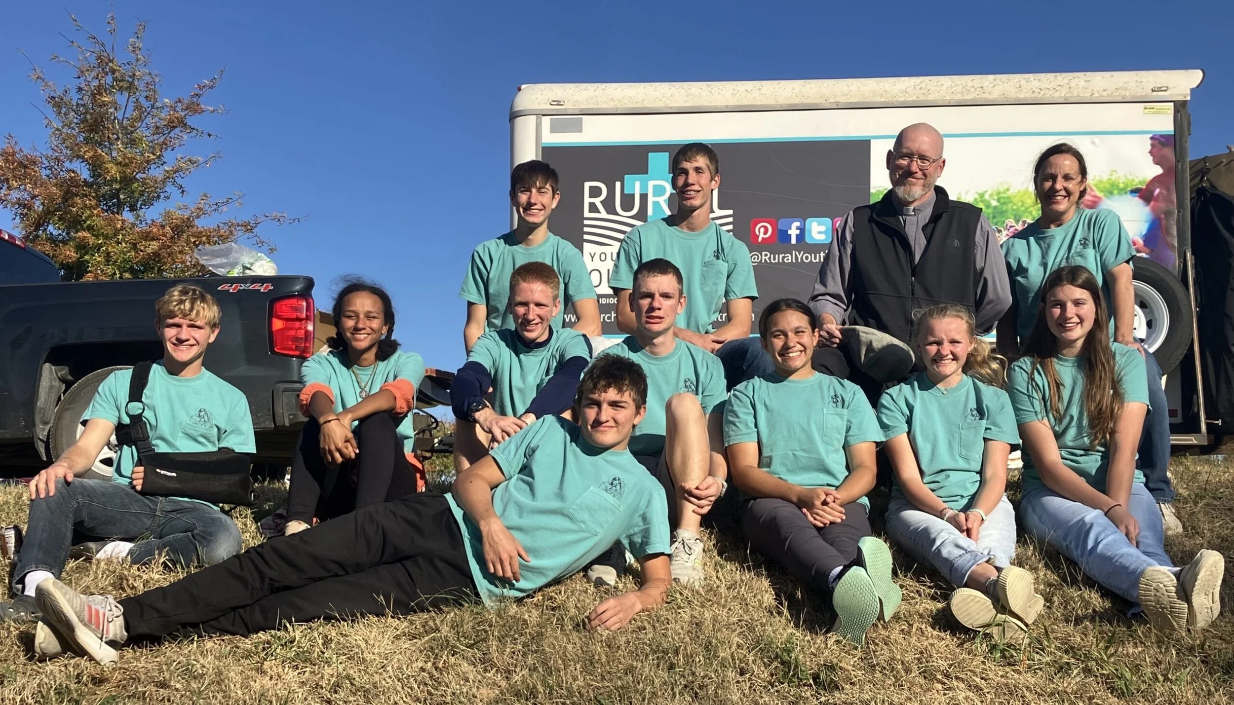 Group of young people with two adults posing on grass in front of a vehicle with a promotional banner for Rural Youth. Everyone is smiling, some sitting, some standing, outdoors on a sunny day.