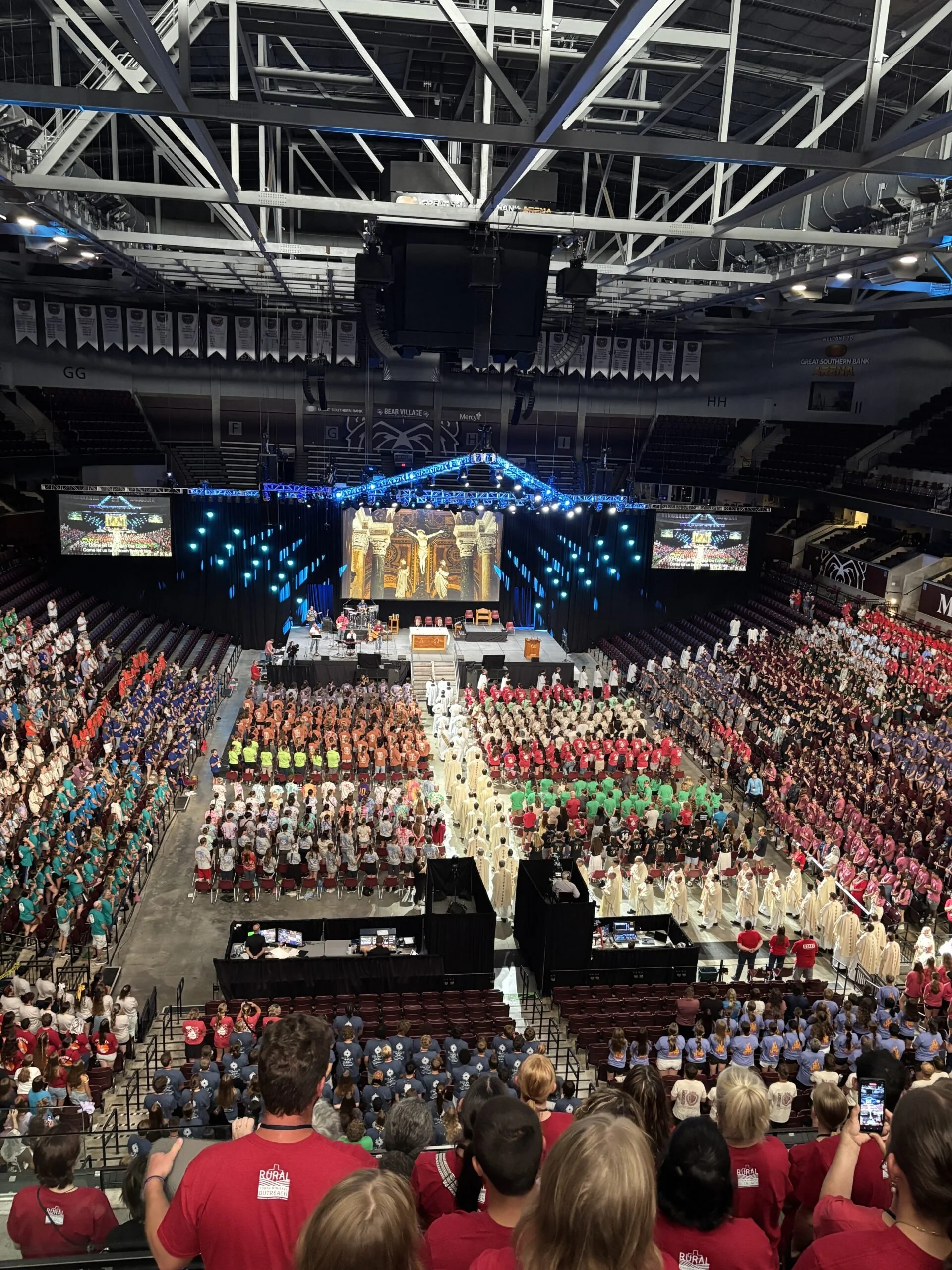 View of a large indoor event with many attendees seated and standing, with a stage at the front featuring a backdrop and decorations, inside a sports arena or convention center.