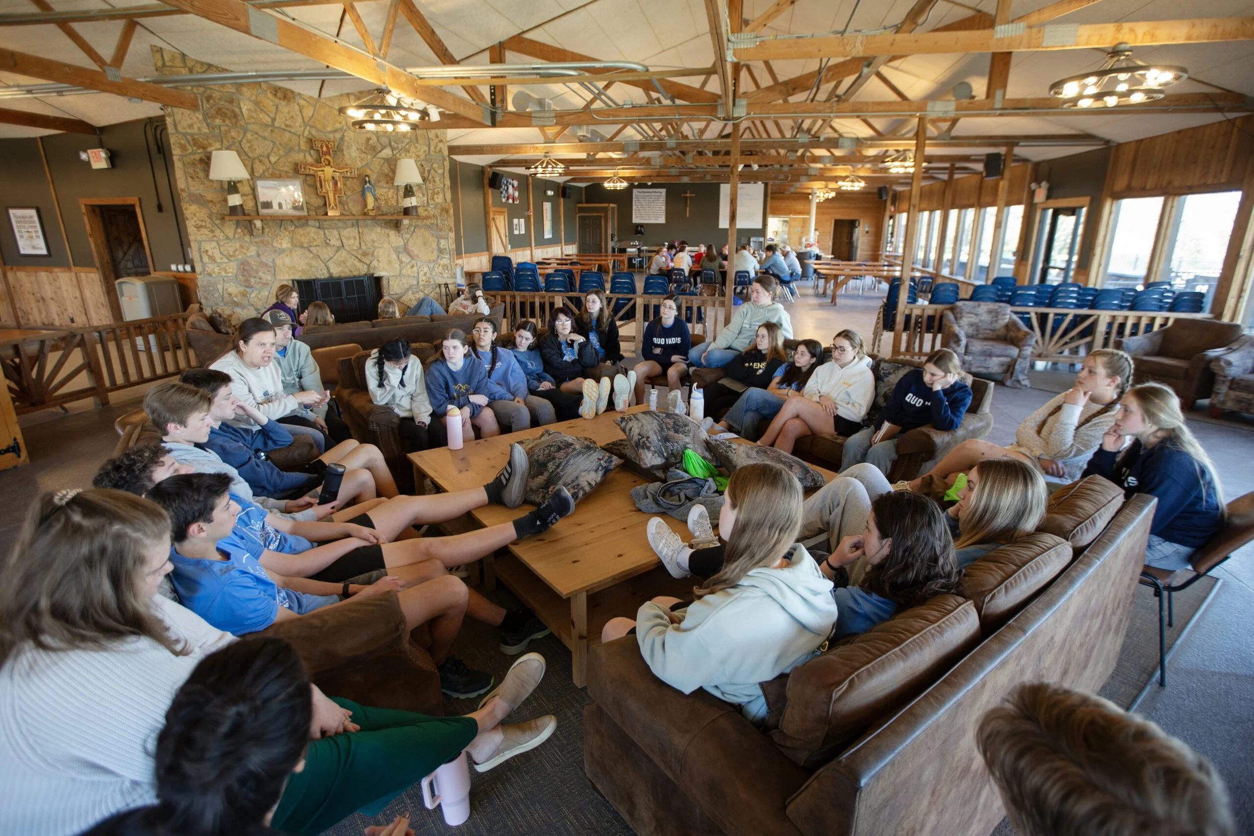 A group of teenagers sitting on couches and chairs in a rustic indoor space, possibly a lodge or retreat center, engaged in a discussion or listening to a presentation. There are large windows on one side, a stone fireplace with religious decorations