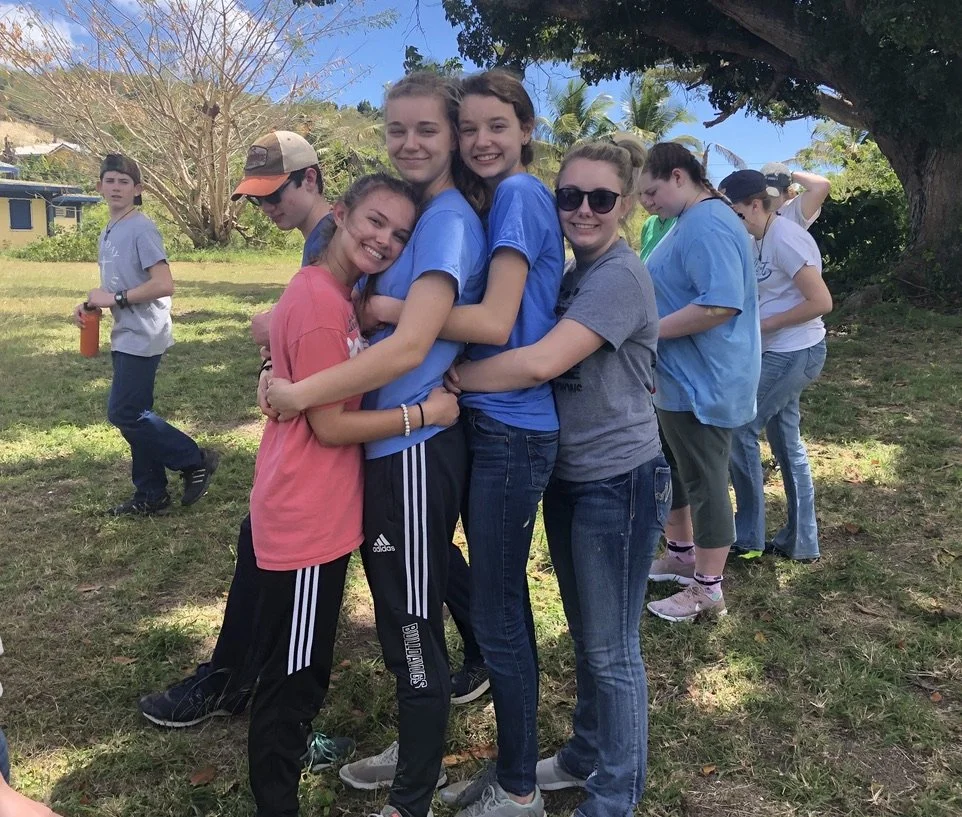 Group of eight smiling teens and preteens hugging each other outdoors under a large tree, with others standing nearby, in a park or yard with green grass and trees.