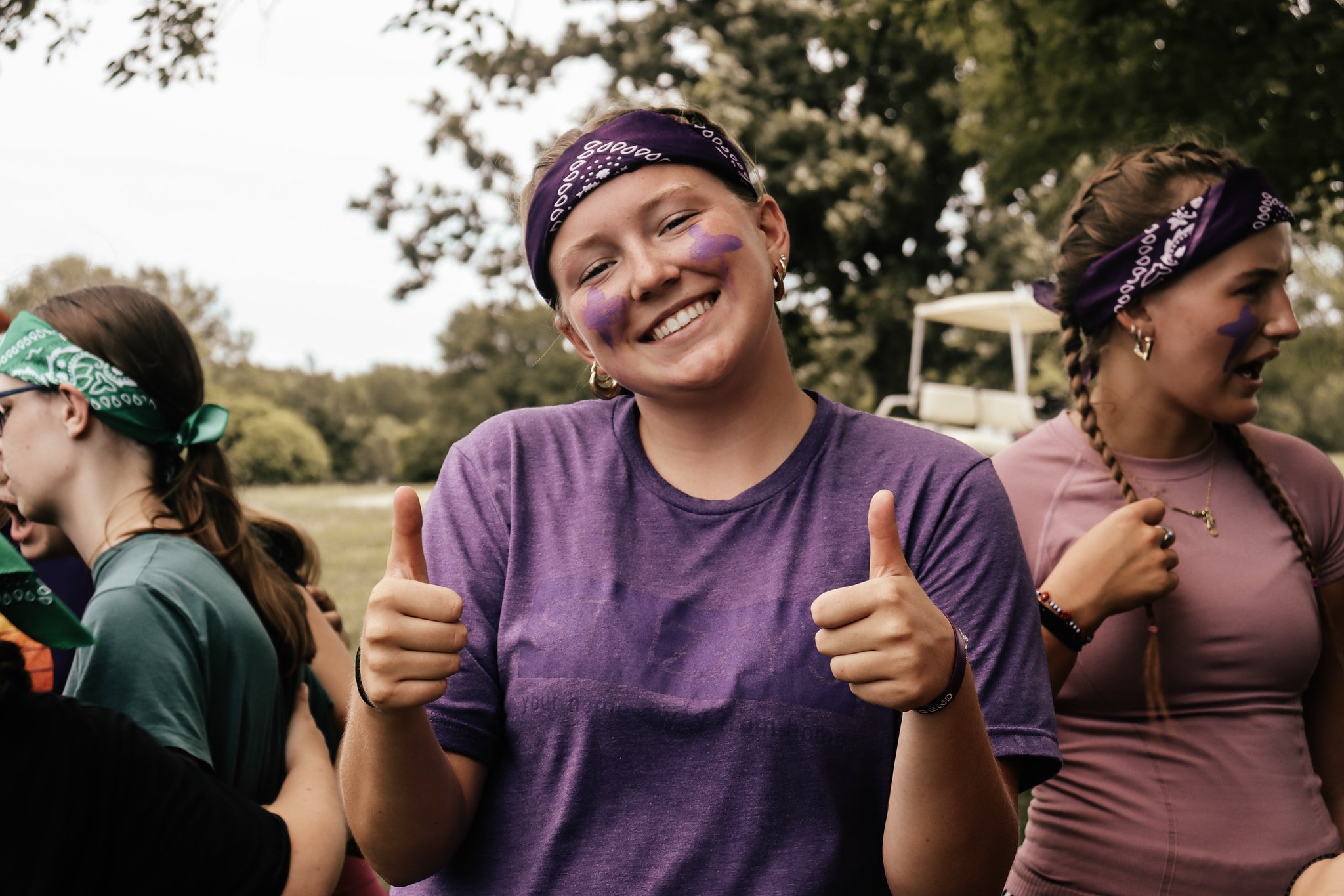 Young woman smiling and giving thumbs up during outdoor event, wearing purple shirt, purple face paint, and purple headband, with people around her.