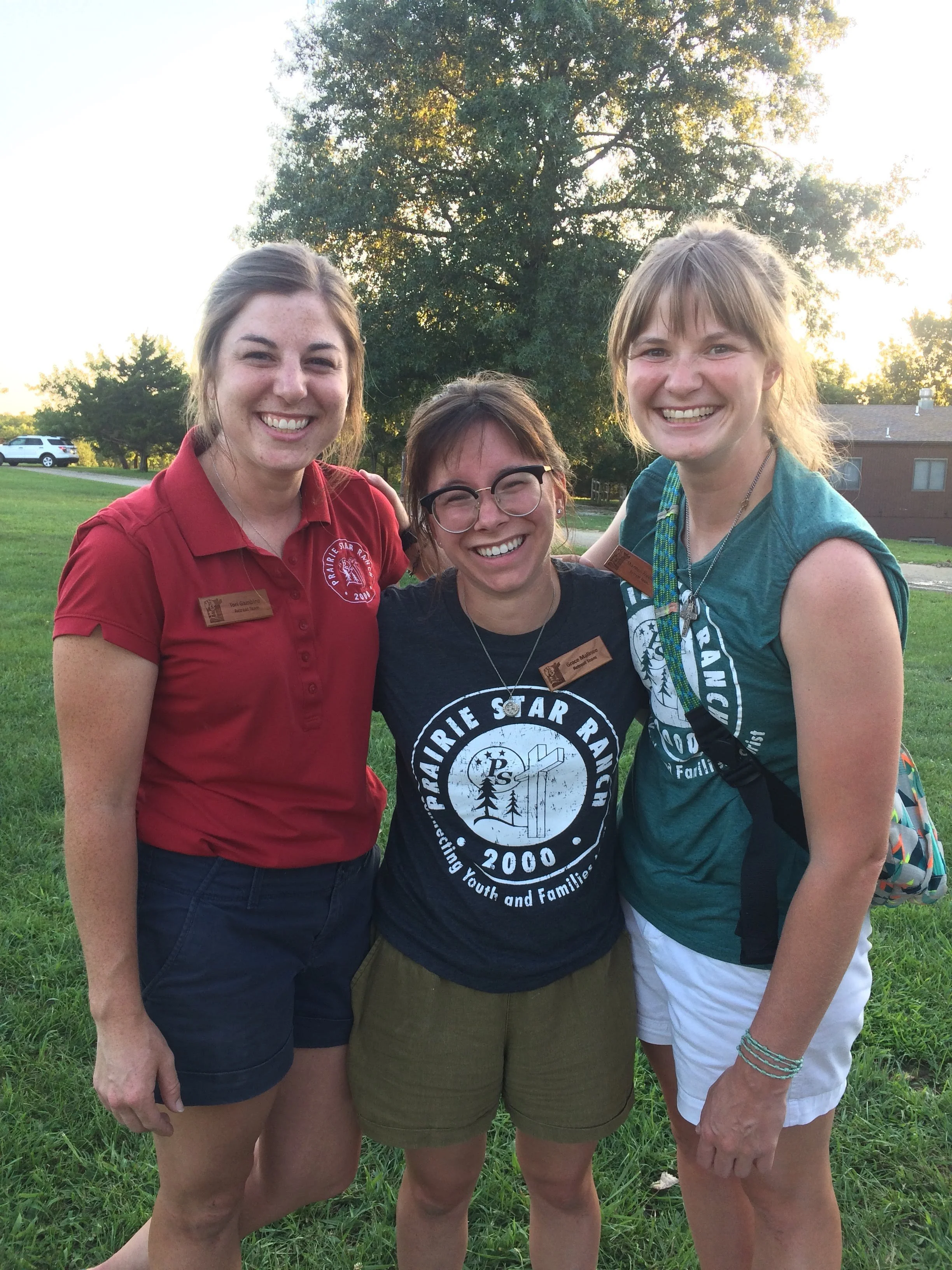 Three young women smiling and standing close together outdoors on a grassy area during daytime, with trees, a house, and a car in the background.