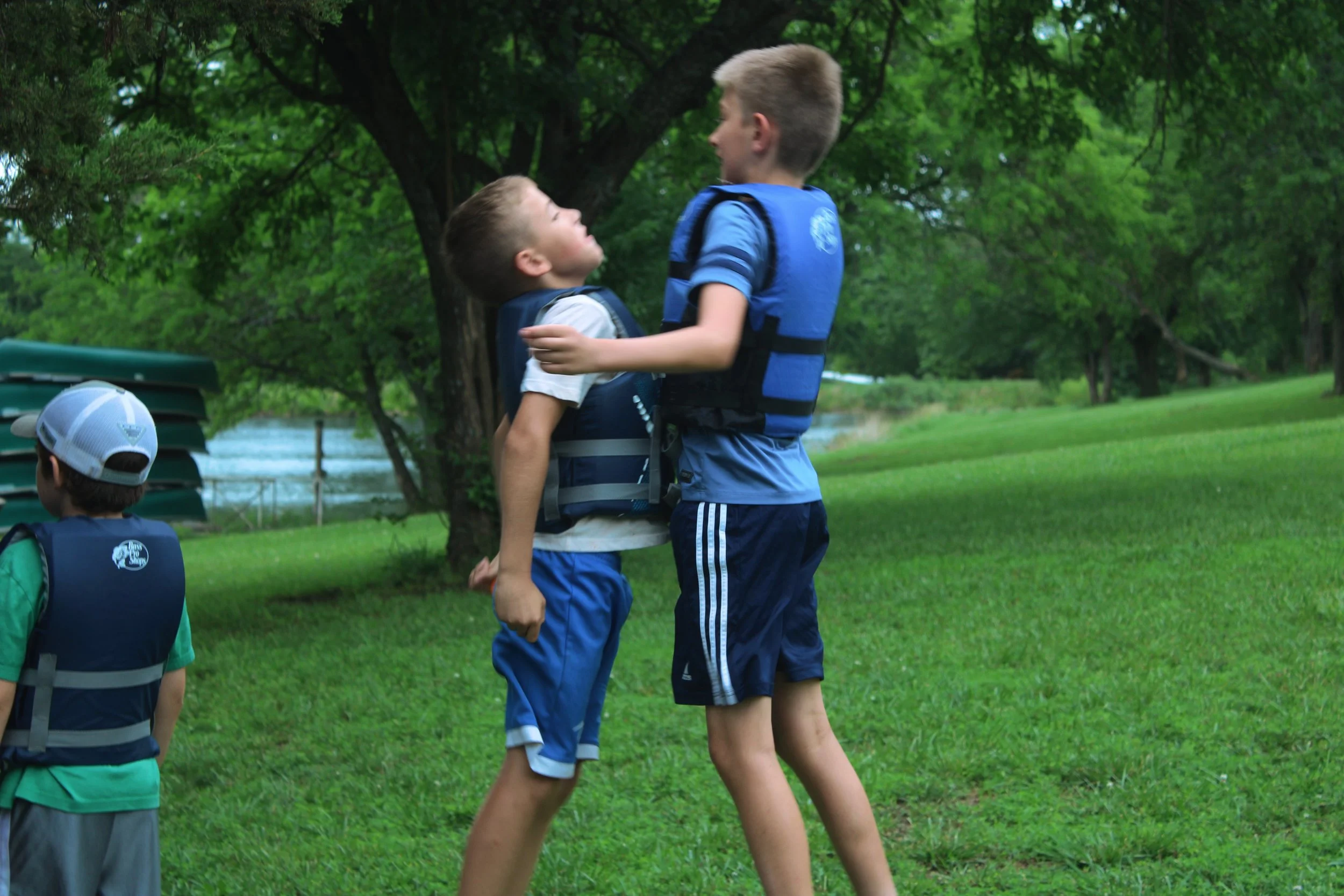 Two boys hugging outdoors near a large tree on a grassy area, with children wearing backpacks nearby, during daytime.