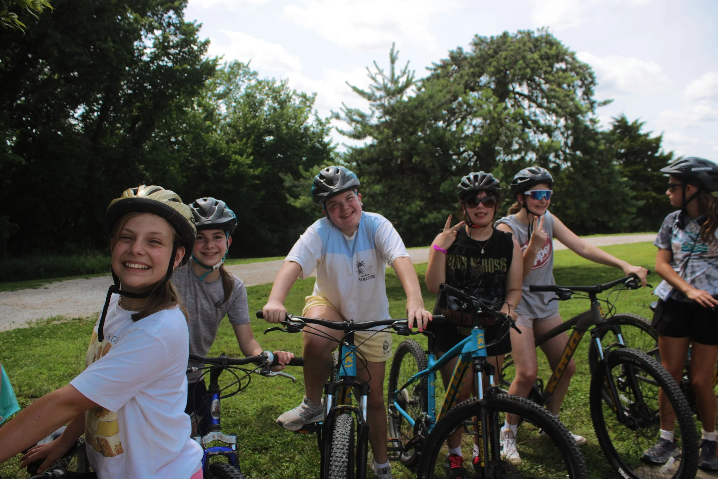 Group of children wearing bicycle helmets and smiling, with some making peace signs, standing with their bikes outdoors in a park-like setting with trees and a blue sky in the background.