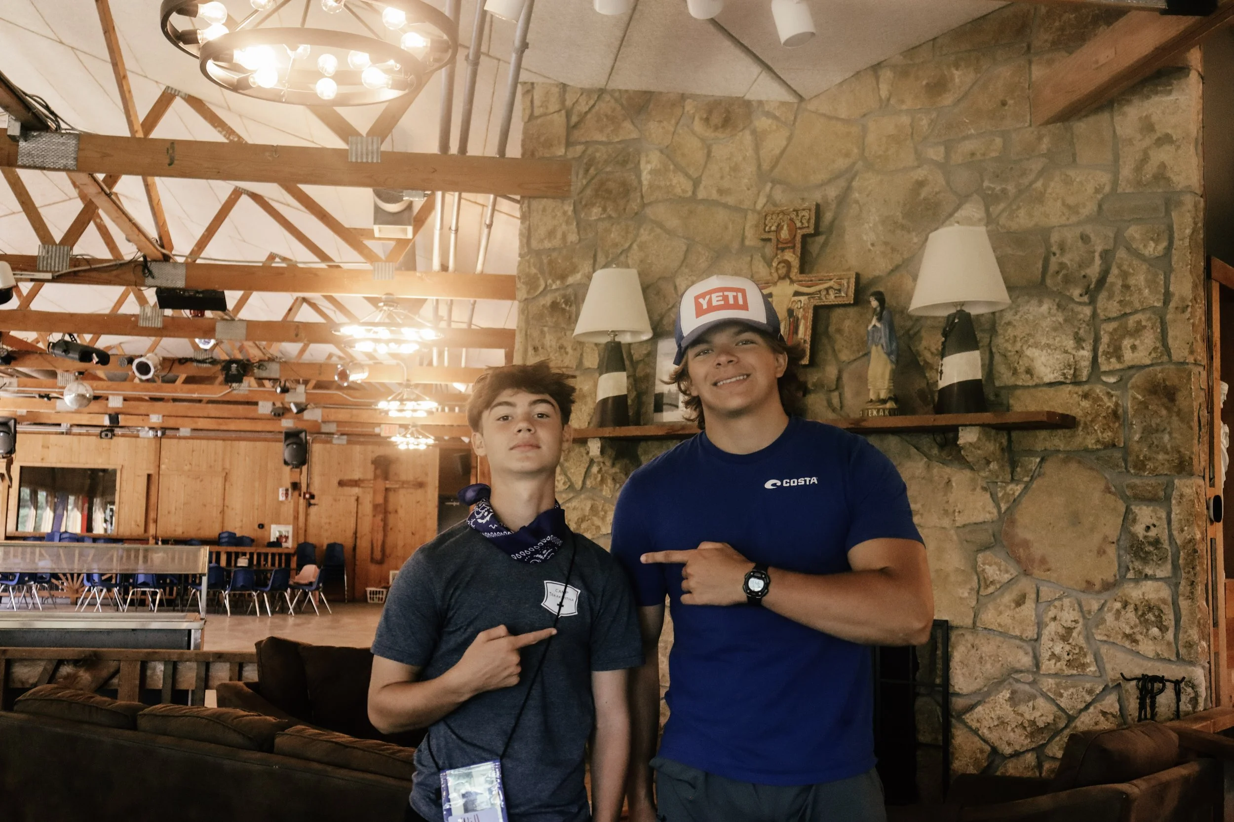 Two young men standing together in a rustic indoor setting with a stone fireplace, wooden beams, and decorative lamps. One is pointing at the other, who is wearing a YETI cap and a blue t-shirt.