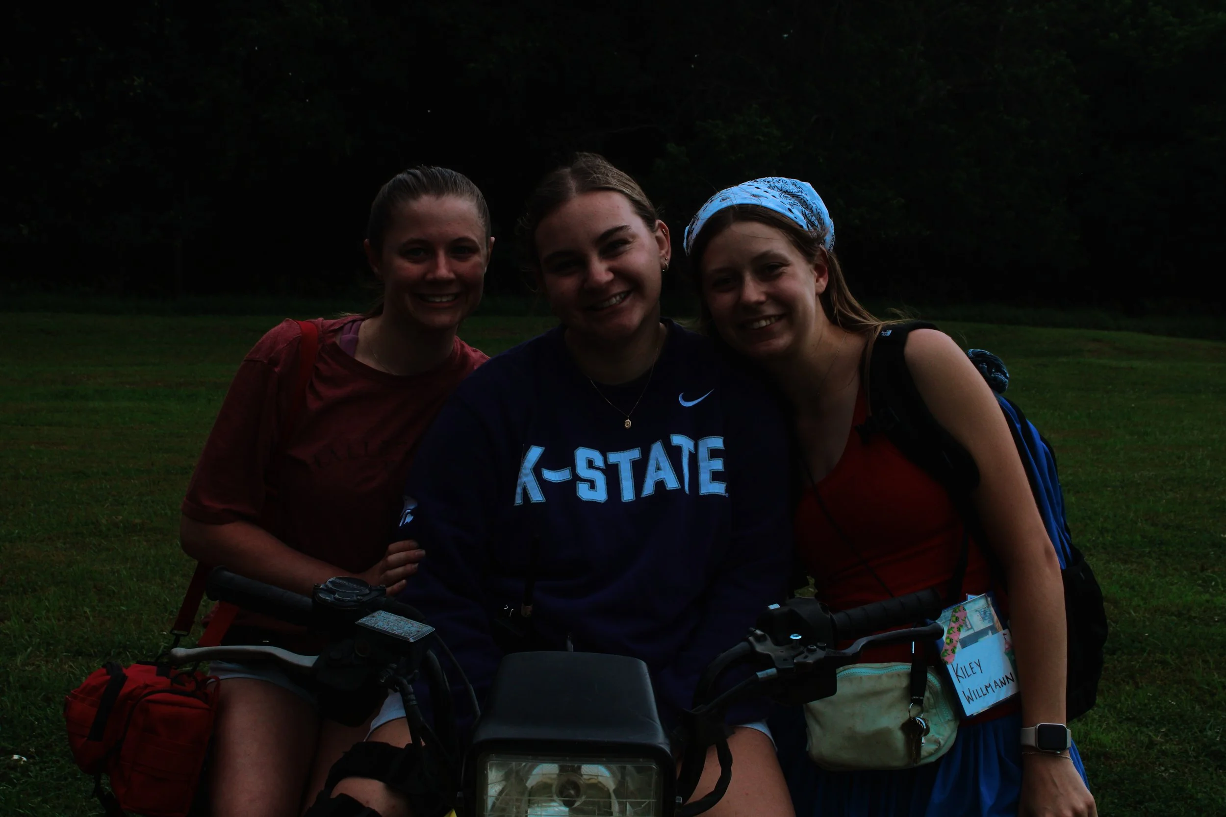 Three young women standing outdoors at dusk, smiling, with dark trees in the background. One woman wears a blue bandana, another a red shirt, and the third a dark shirt with 'K-STATE' written on it.