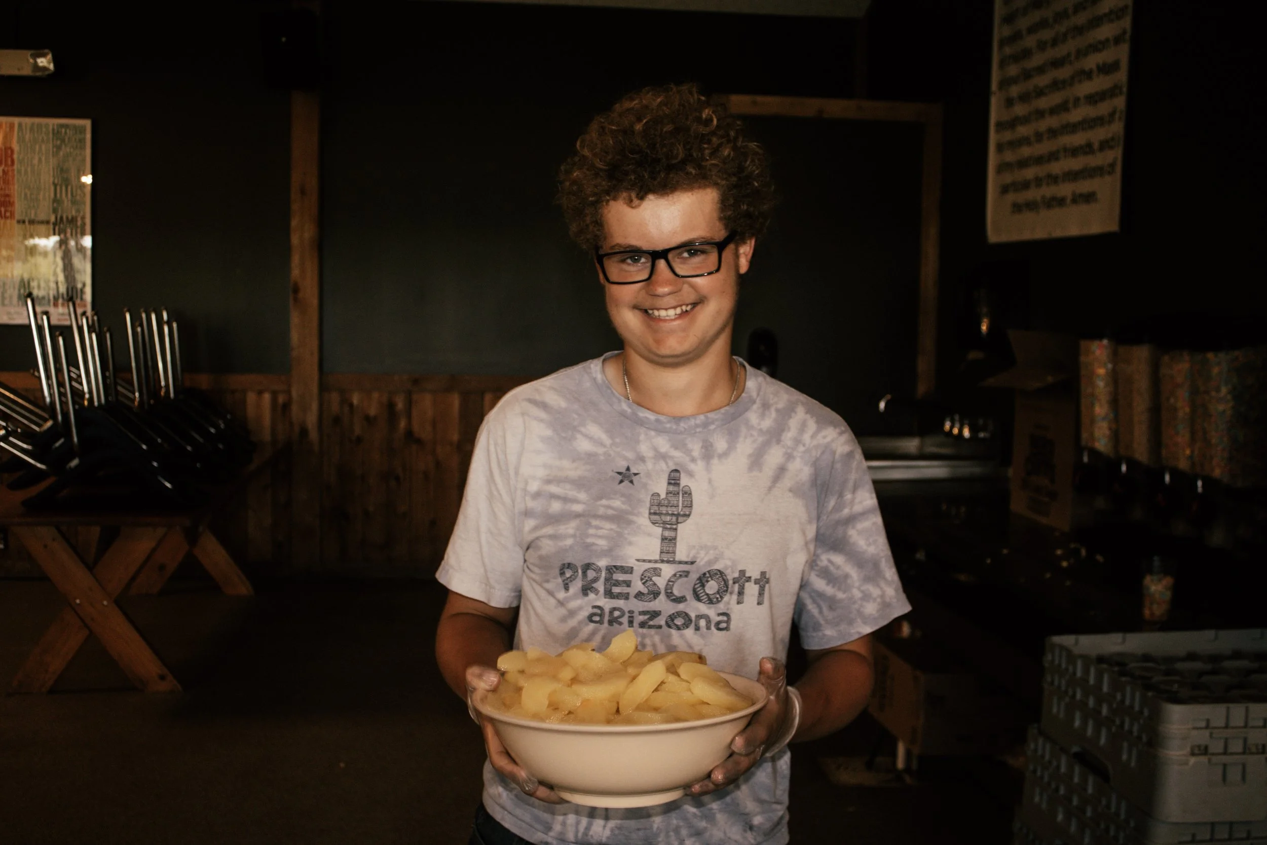 A young man with curly hair and glasses smiling, holding a bowl of sliced potatoes in a dimly lit indoor setting.