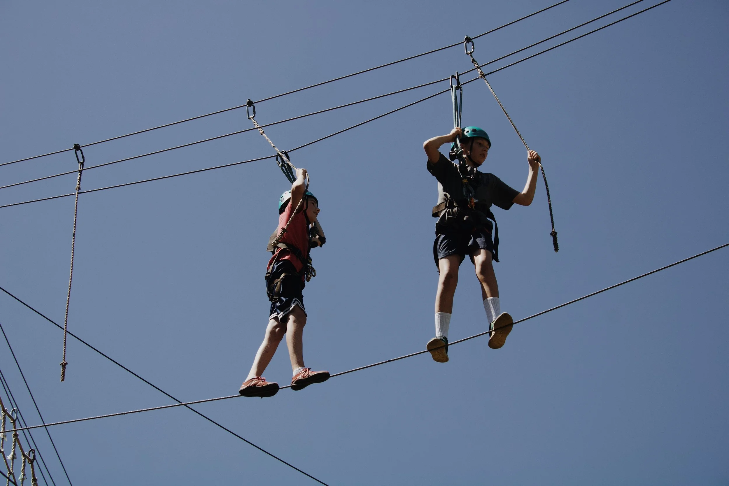 Two children wearing helmets and harnesses walking on a high ropes course or obstacle bridge suspended between cables against a clear blue sky.