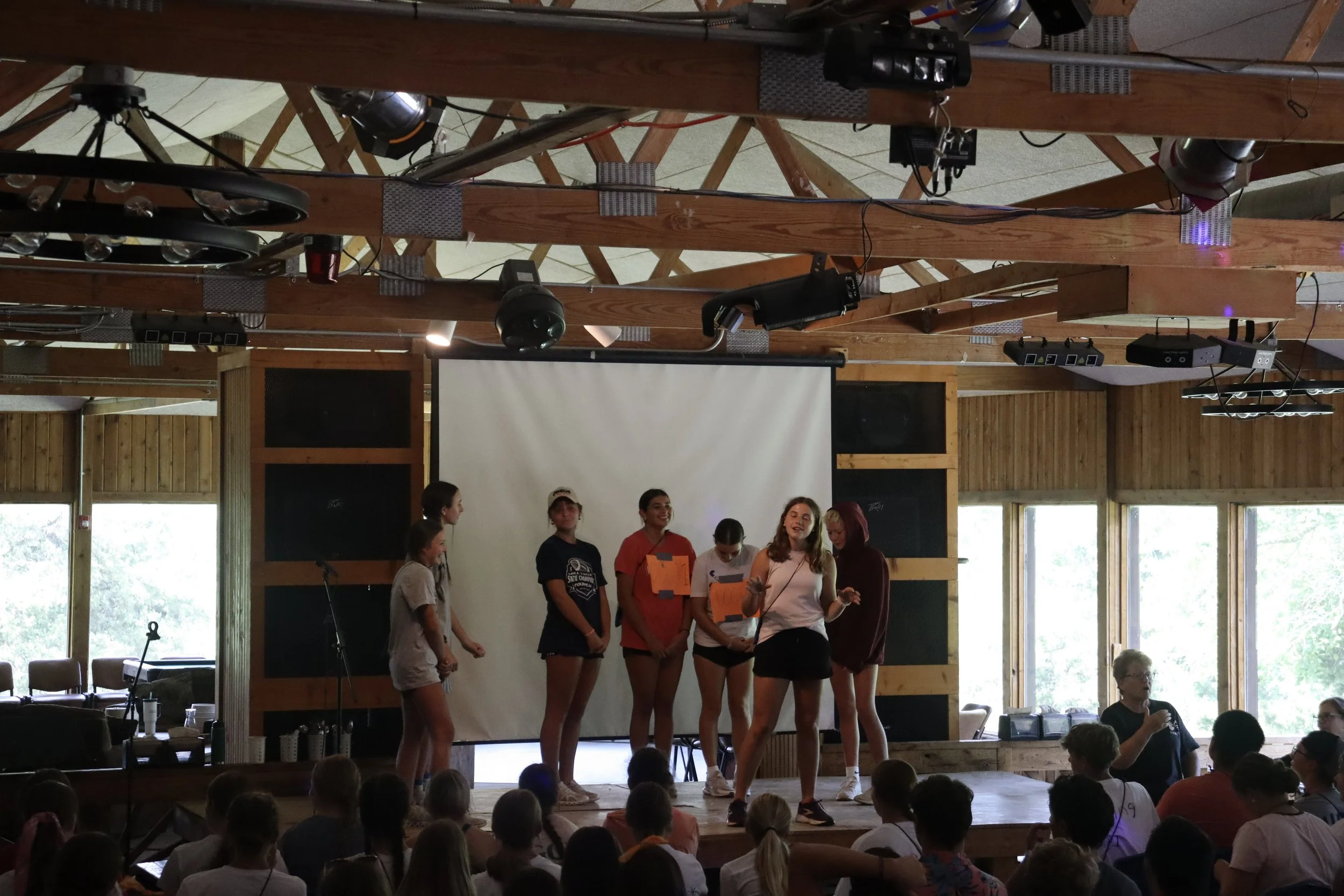 Group of young women standing on stage in a wooden hall. Audience seated in front, some visible at the bottom of the image. Stage has a white projector screen and lighting equipment hanging from the ceiling.