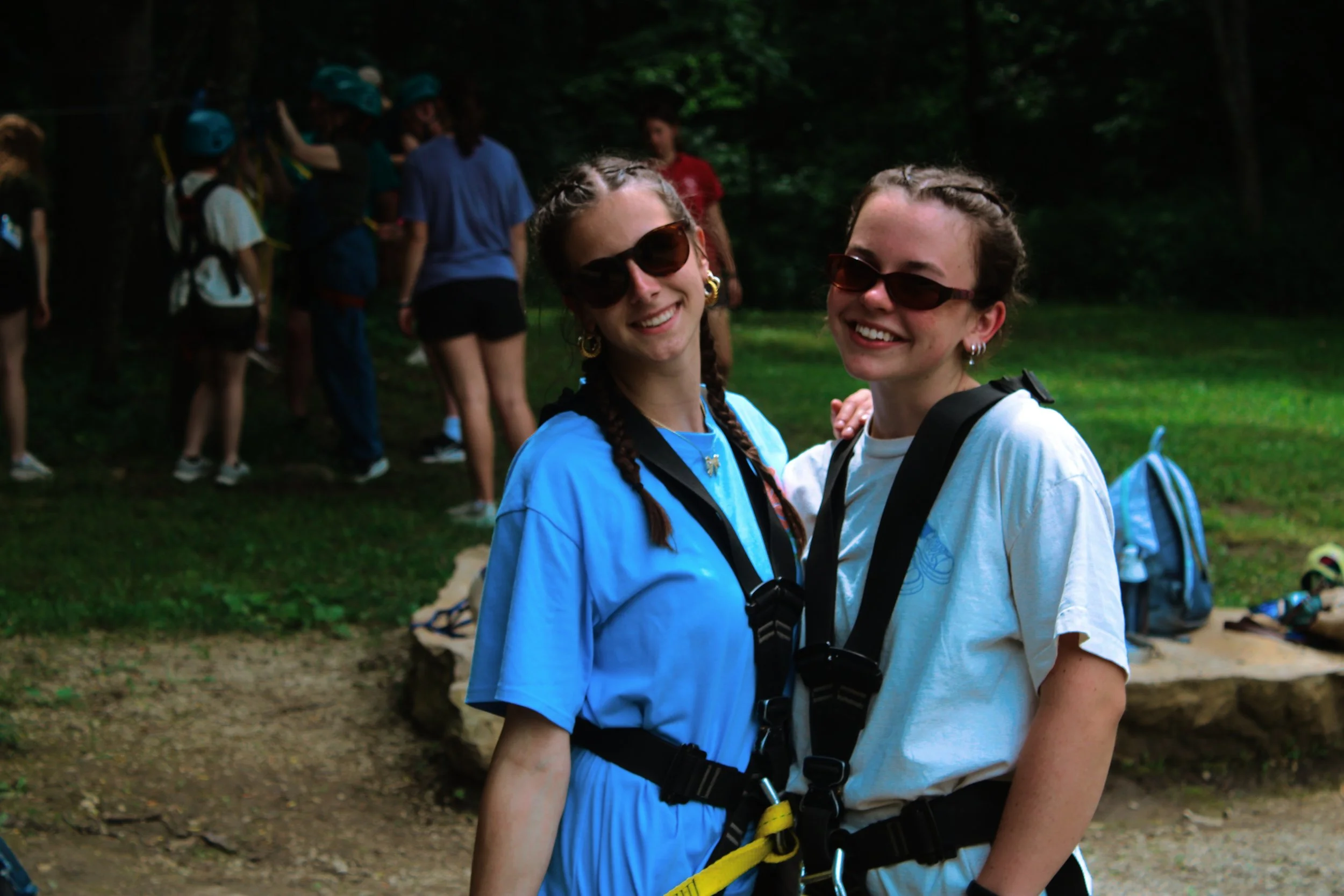 Two young women with braided hair and sunglasses smiling outdoors, wearing harnesses, with a group of people in safety gear in the background in a wooded area.