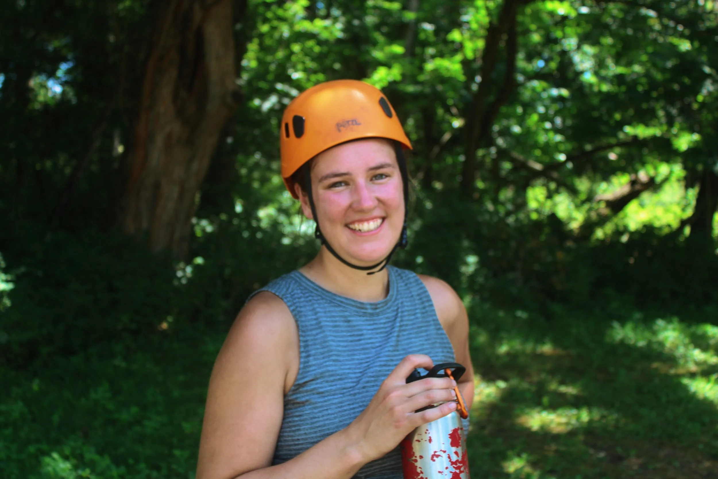 A young woman wearing an orange helmet and a blue sleeveless top smiling outdoors in a wooded area, holding a drinks bottle.