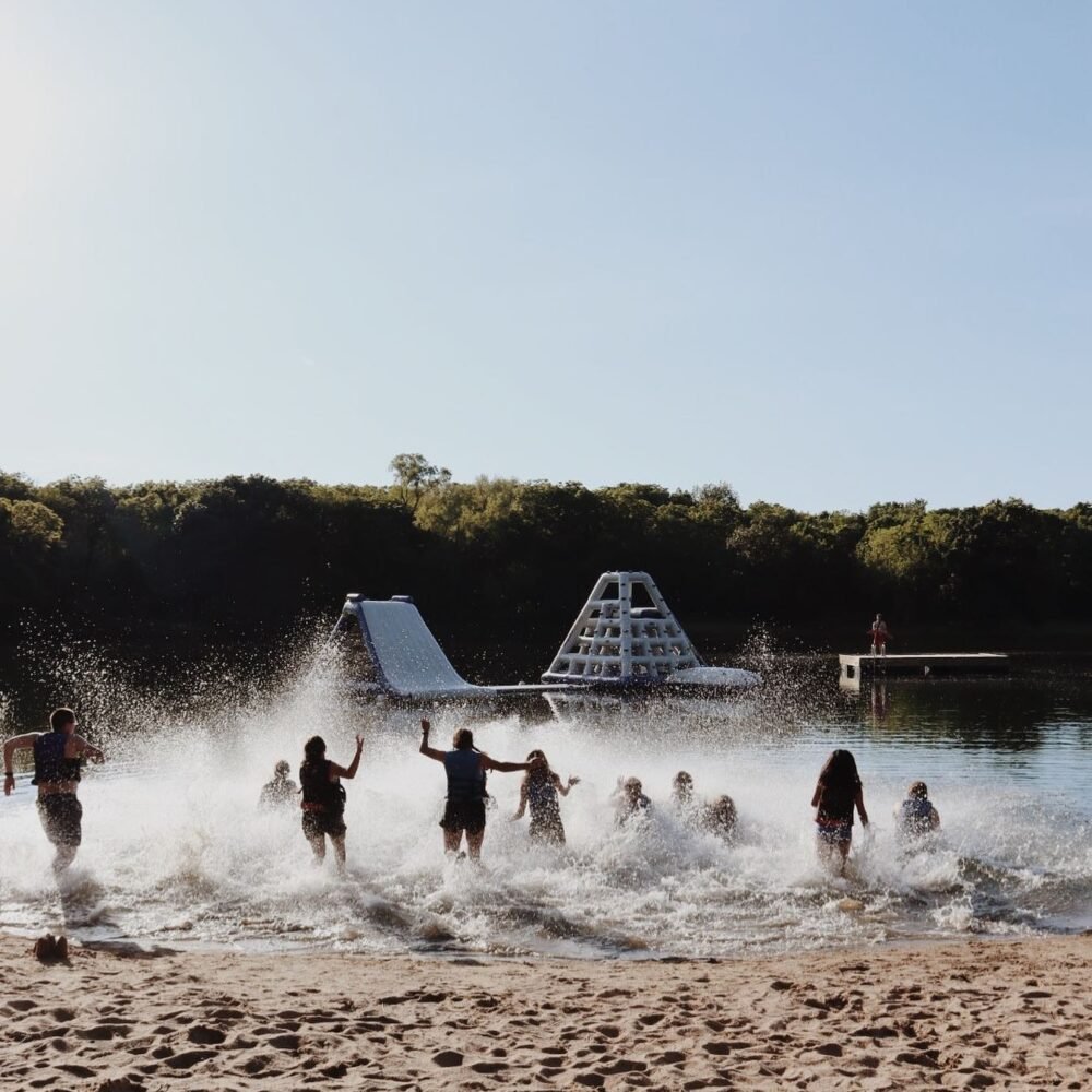People playing in shallow lake water near a children's water park with a slide and climbing structure