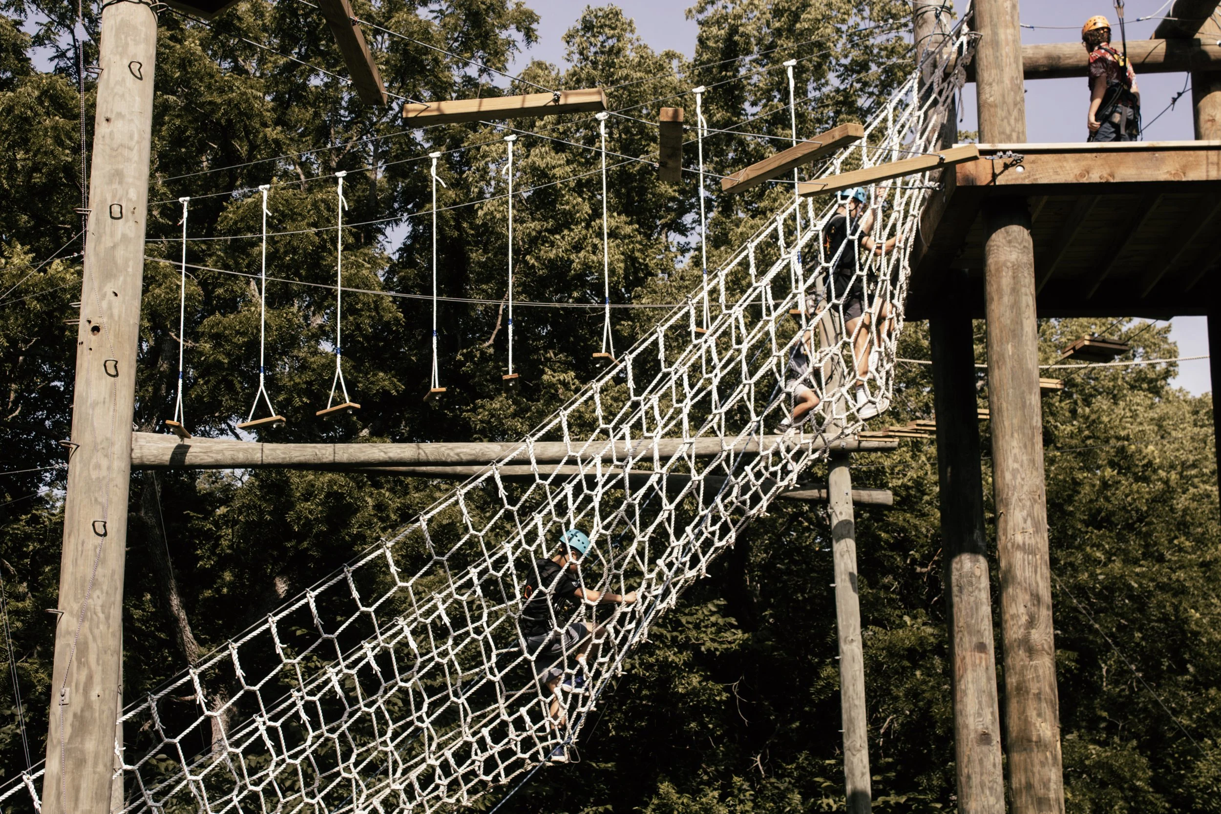 Children wearing helmets climbing a rope bridge at an outdoor adventure park during daytime, surrounded by tall trees.