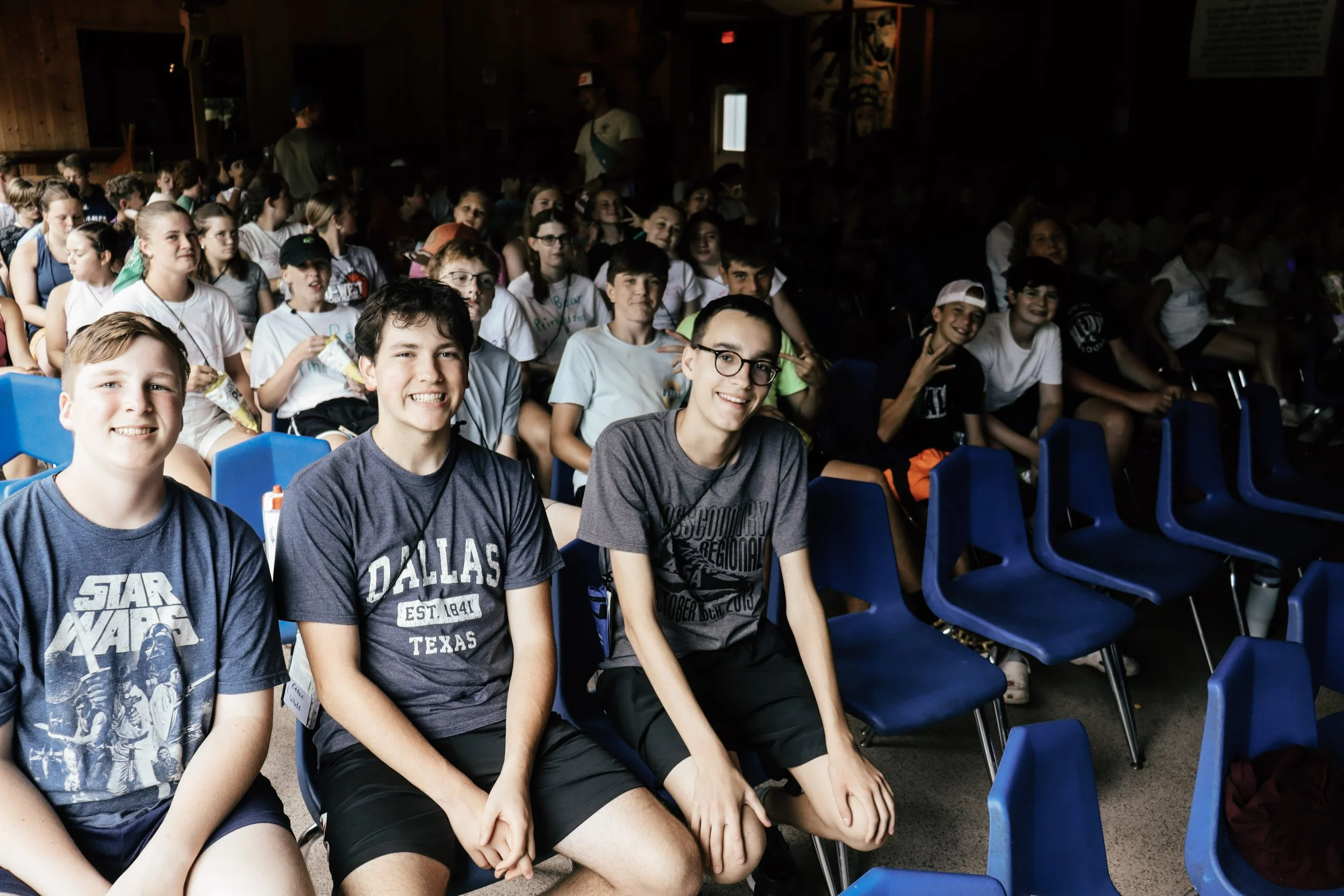 A group of young students sitting in blue plastic chairs inside a dimly lit room, smiling and posing for the camera. Some students are wearing casual t-shirts, and a few are making peace signs.