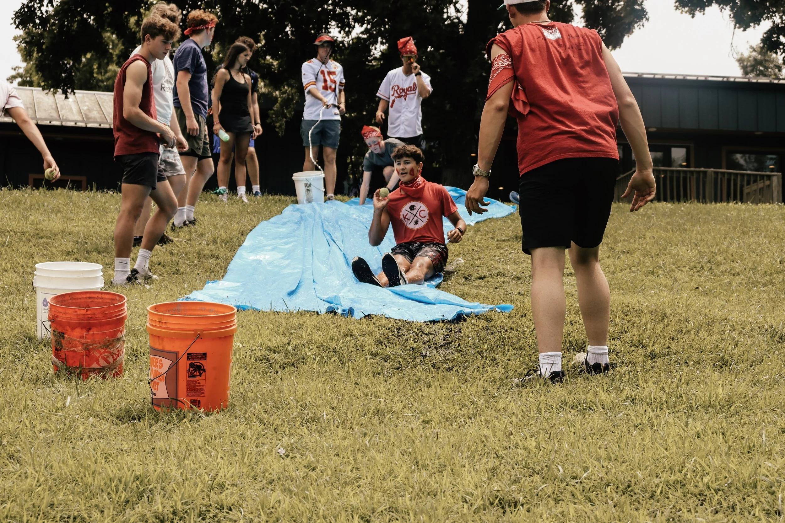 Group of young people at an outdoor water activity, with one person sitting on a blue tarp holding a ball, others standing around, and buckets of water in the foreground.