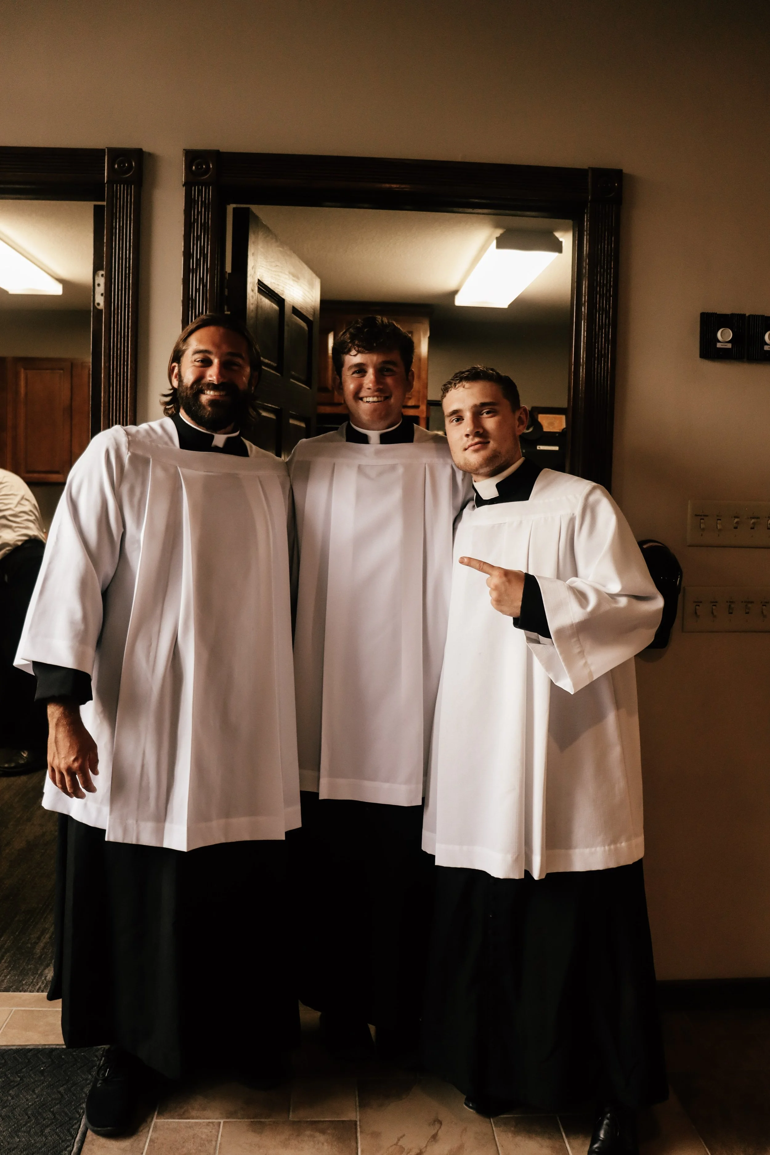 Three men wearing traditional academic robes and clerical collars standing in front of a mirror in a room, smiling at the camera.