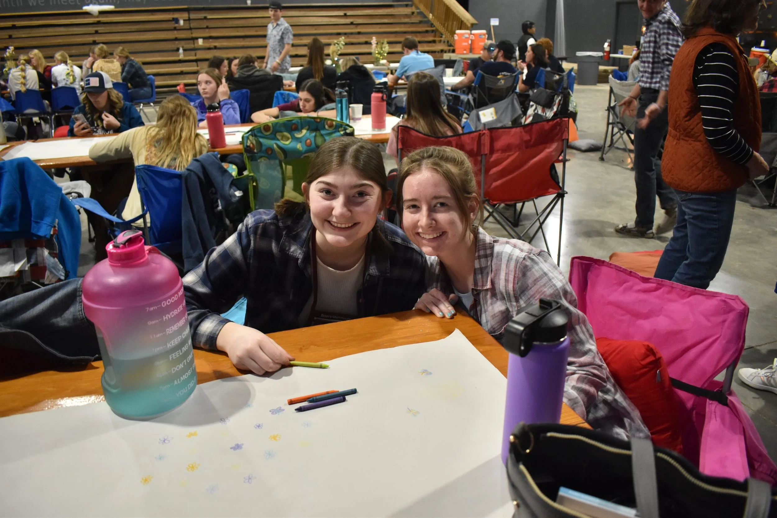 Two young girls smiling and sitting at a table with colored markers and artwork in progress, surrounded by backpacks and water bottles, in a busy indoor event space with many people seated at tables and engaging in activities.