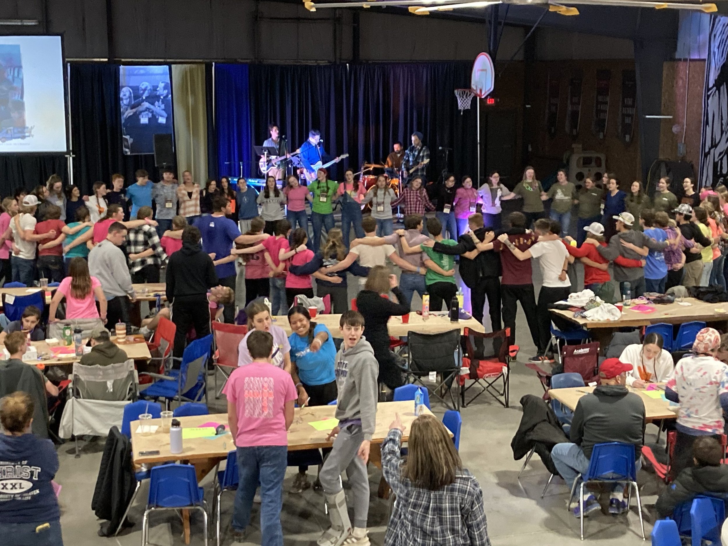 Large gathering of people in a gymnasium with a stage, where a band is performing. People in the audience are standing in a circle holding hands, some seated at tables, participating in a group activity. The gym features a basketball hoop and screens