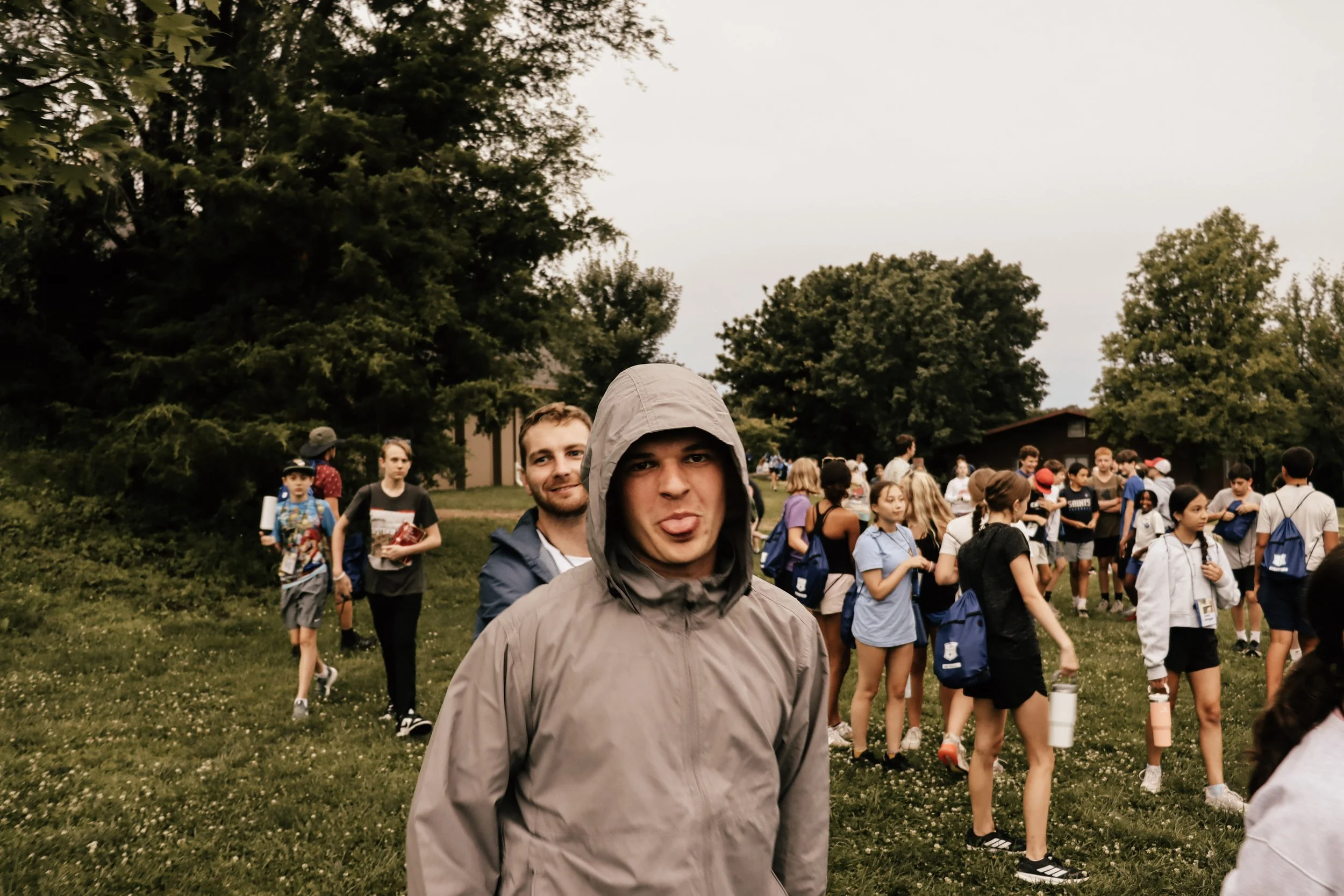 A group of children and teenagers gathered outdoors on a grassy area with trees in the background, some wearing backpacks, and two young men in the foreground, one with a hood and the other sticking out his tongue.