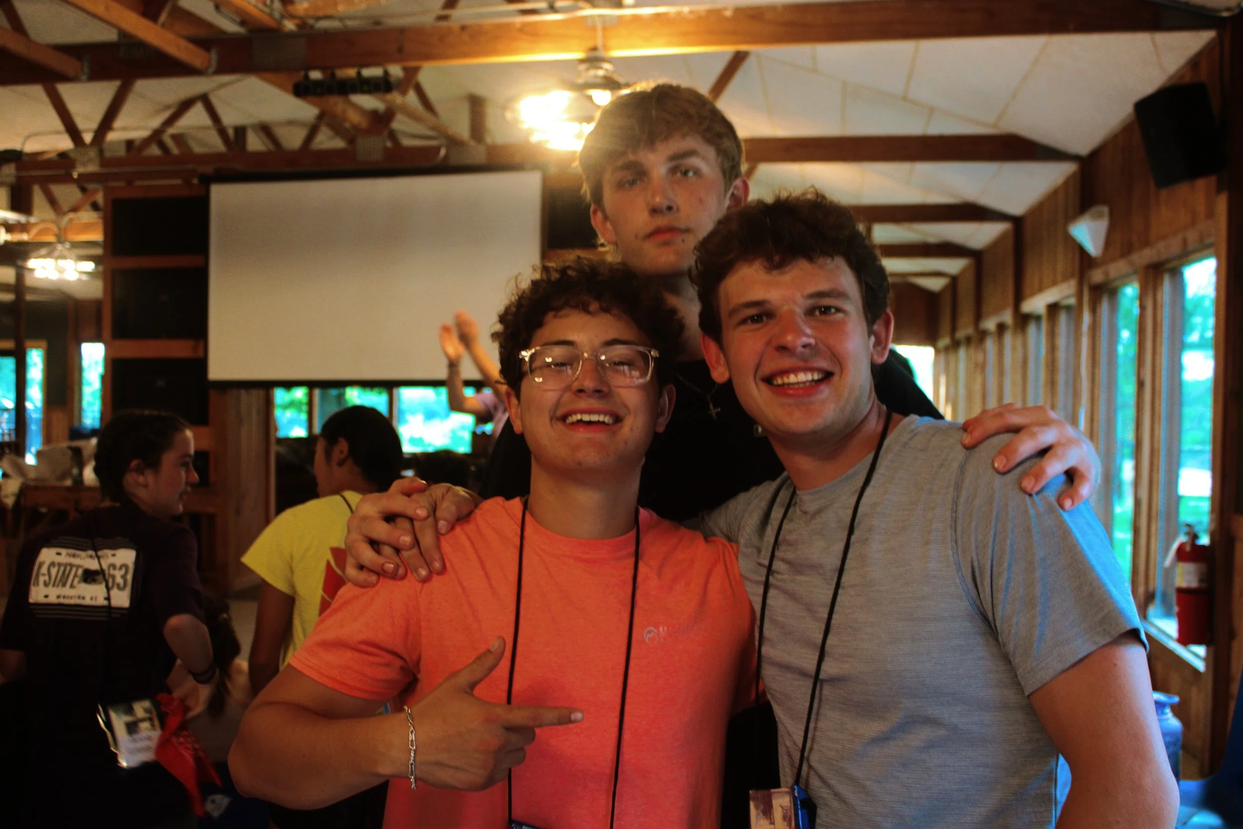 Three young men are standing close together indoors at a gathering or event, smiling at the camera. The environment features wooden walls and a ceiling with exposed wooden beams and ceiling fans. Other people are present in the background, some engag