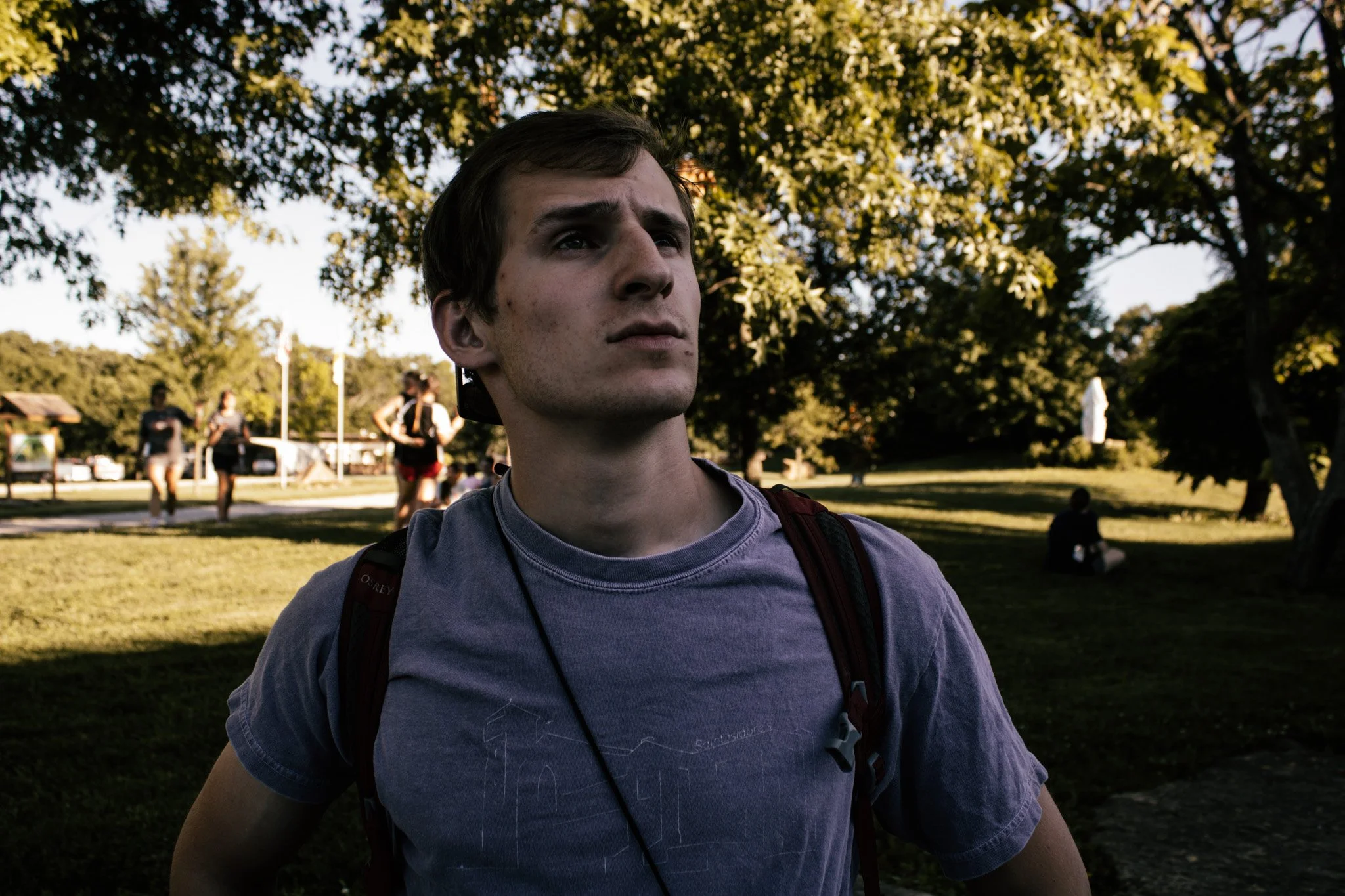 Young man with dark hair and a serious expression, wearing a gray T-shirt and carrying a backpack, standing in a park with others and trees in the background.