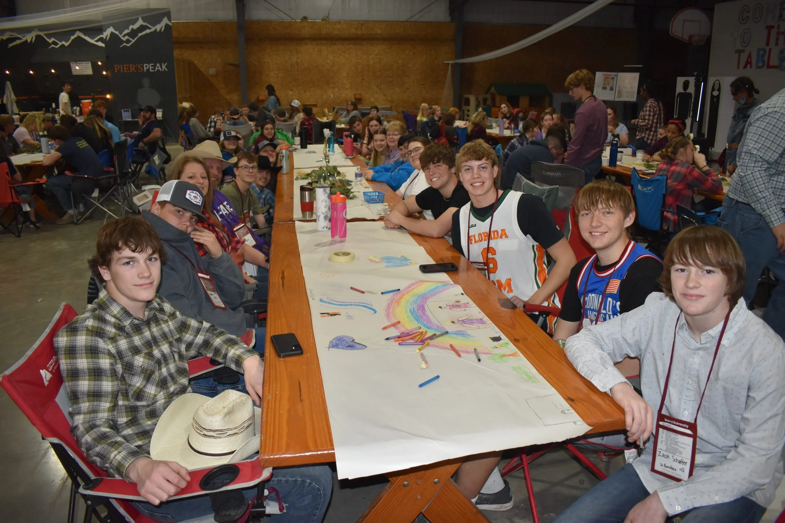 A group of teenagers sitting at a long table in an indoor community center or sports hall, smiling at the camera. The table has drawings and art supplies. Some attendees are in casual clothes, and one is in a basketball jersey. There is a mural of mo