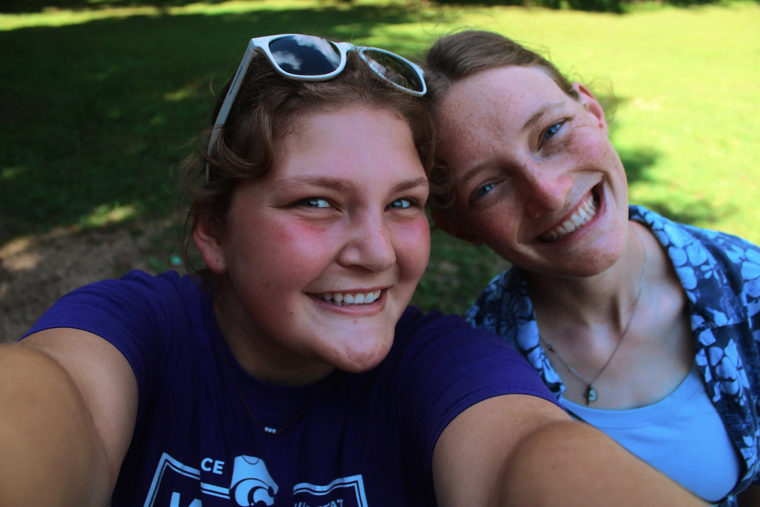 Two teenagers, a girl and a boy, smiling and taking a selfie outdoors on a sunny day, with green trees and grass in the background.