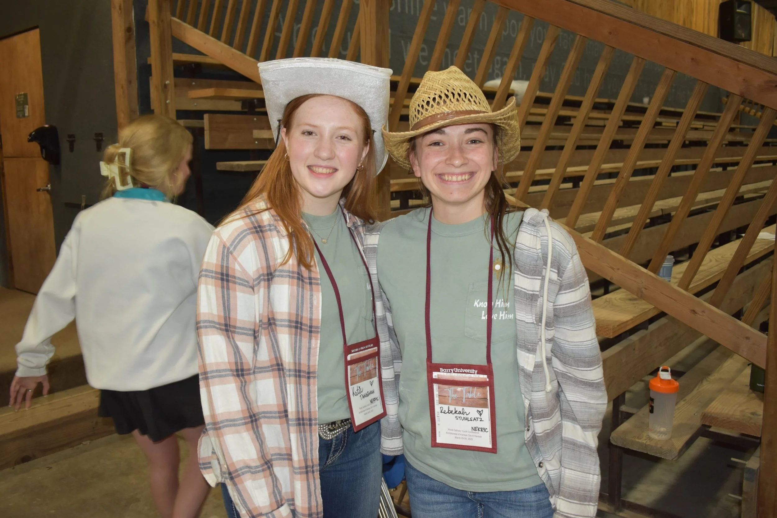 Two young women smiling and wearing cowboy hats at an indoor event with wooden bleachers and a chalkboard in the background.