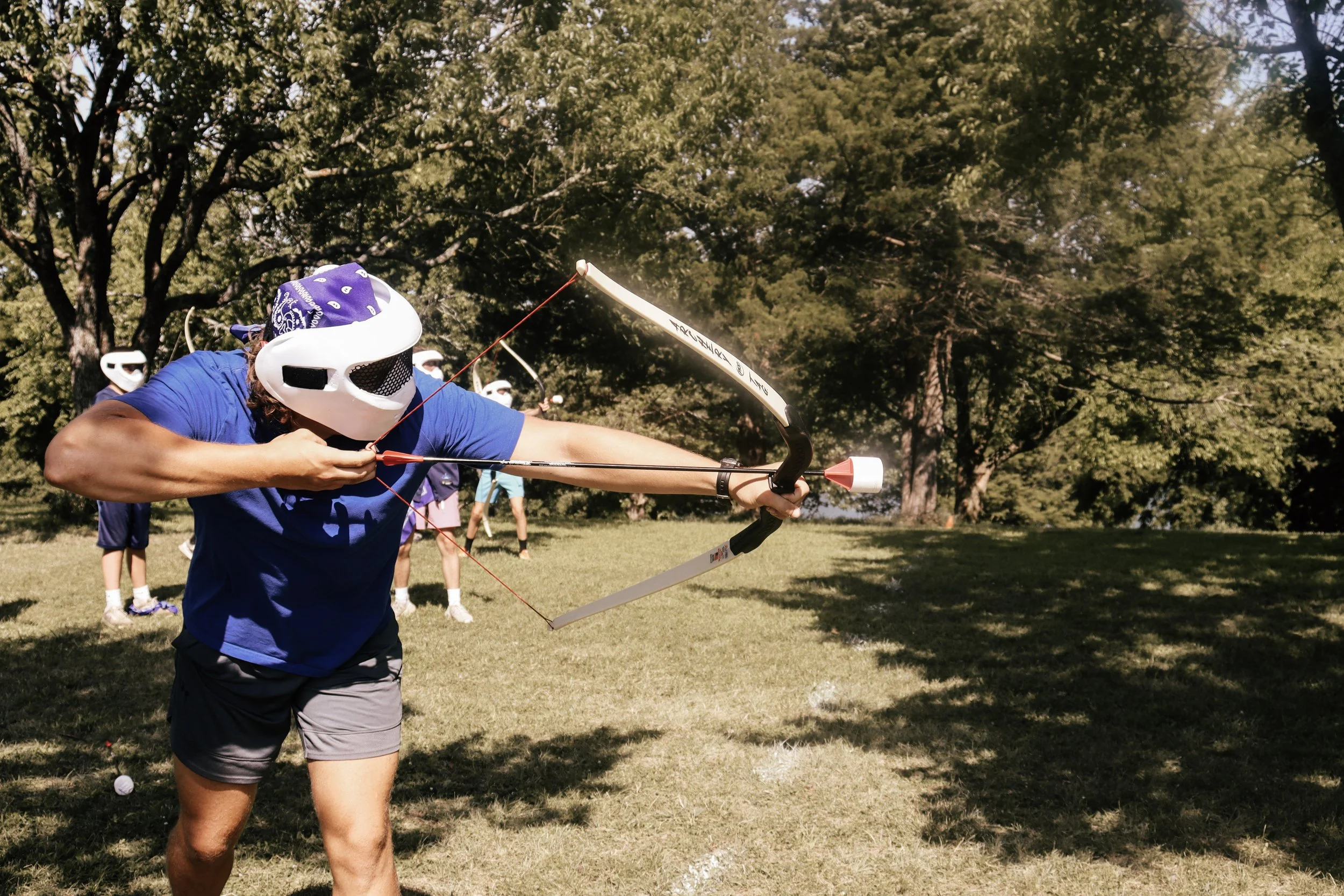 A person wearing a white protective mask and a blue bandana is practicing archery outdoors on a grassy field, surrounded by trees, with several other people in the background also wearing masks.