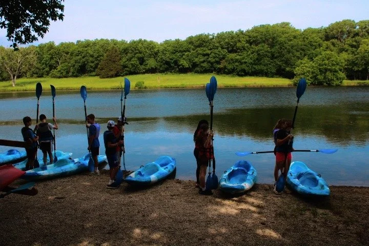 Children with kayaks and paddles by a lake shore during daytime.