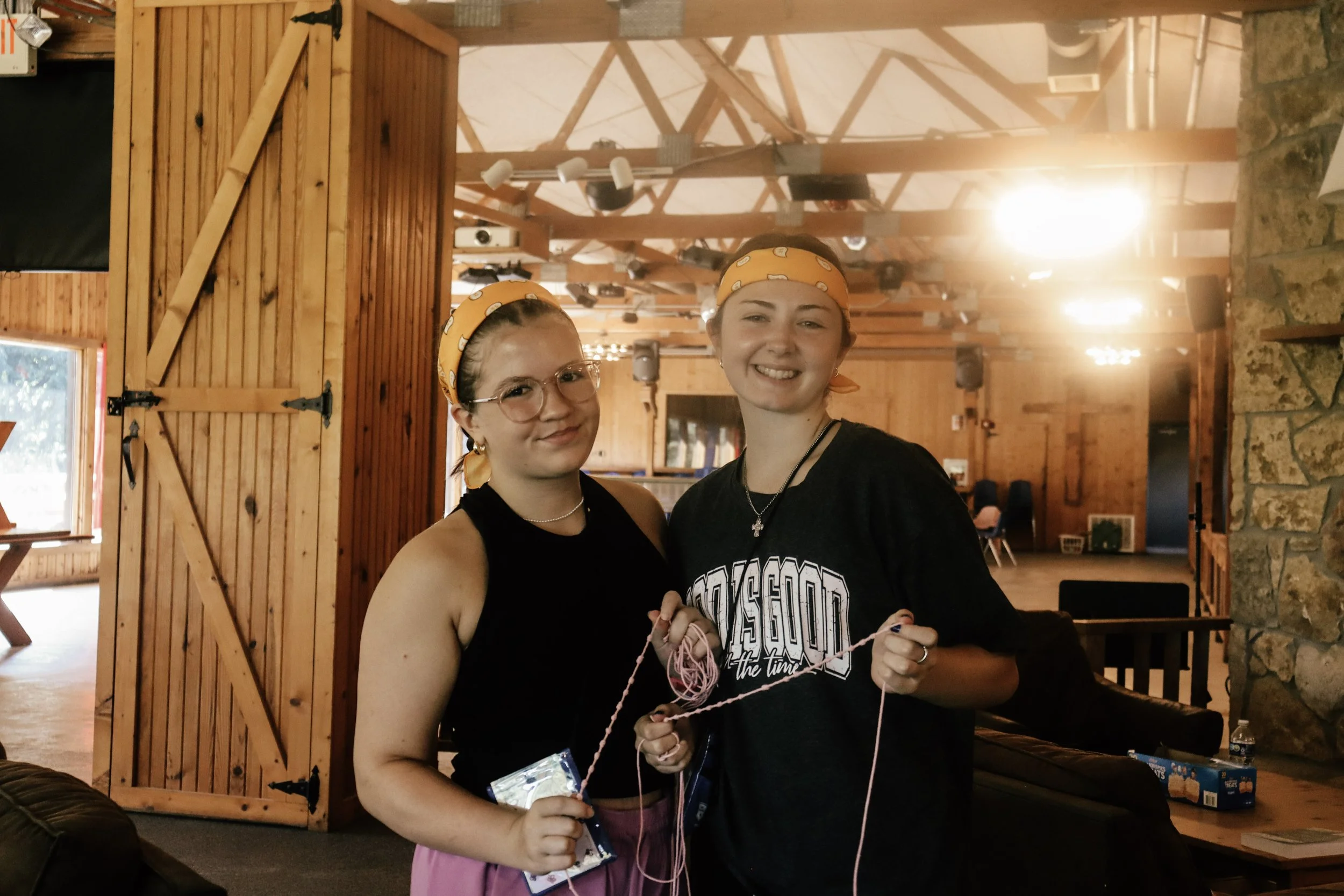 Two young women smiling and standing close together inside a rustic wooden building, wearing yellow bandanas. They are holding knitting yarn and supplies, with the woman on the right wearing a black T-shirt with a graphic design.