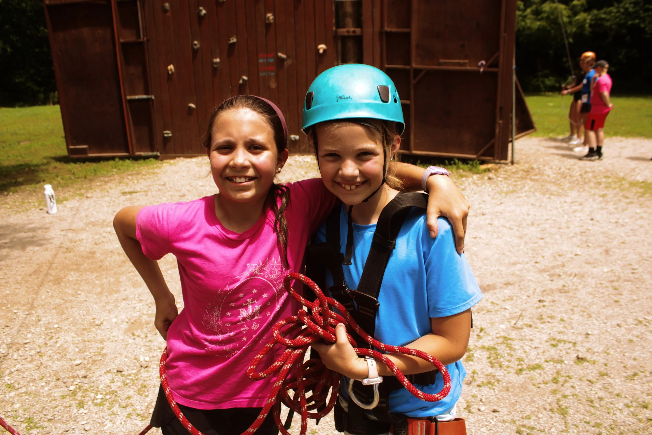 Two smiling young girls standing outdoors at a climbing wall, one wearing a pink shirt and the other wearing a blue helmet and harness, with a group of people in the background.