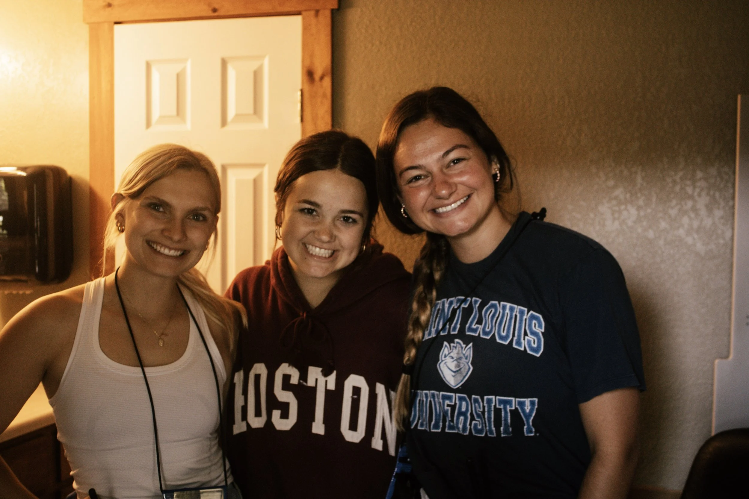 Three young women smiling and posing for a photo indoors, one wearing a white top, another in a maroon hoodie, and the third in a blue university t-shirt with a wolf logo.
