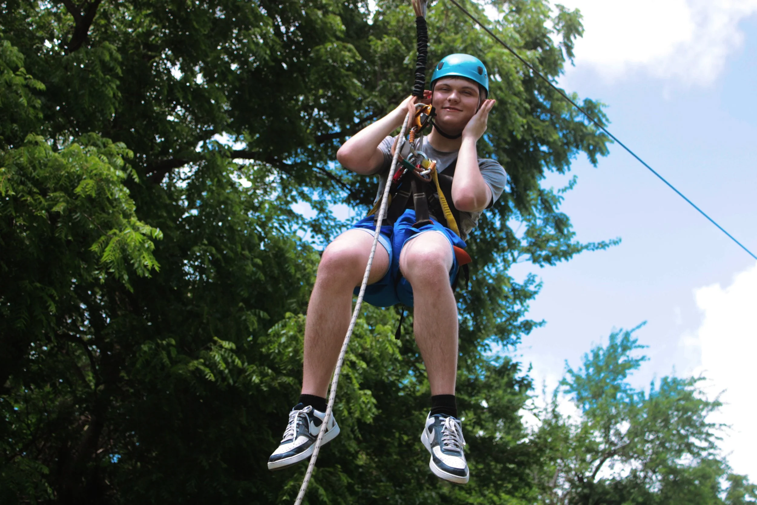 Young man wearing a blue helmet and harness, zip lining through a green wooded area, holding onto the zip line cable with a smile.