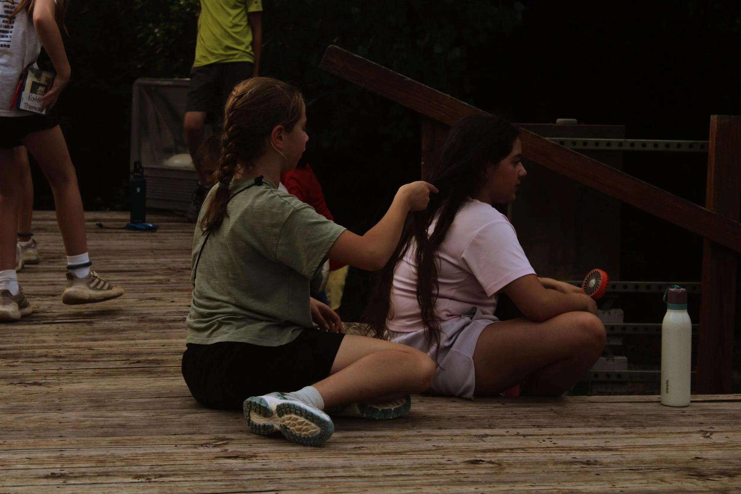A group of young girls are sitting and kneeling on a wooden deck outside, with one girl braiding another girl's hair. In the background, a person is standing and holding a book, and another person is standing near a cooler. Various objects like water