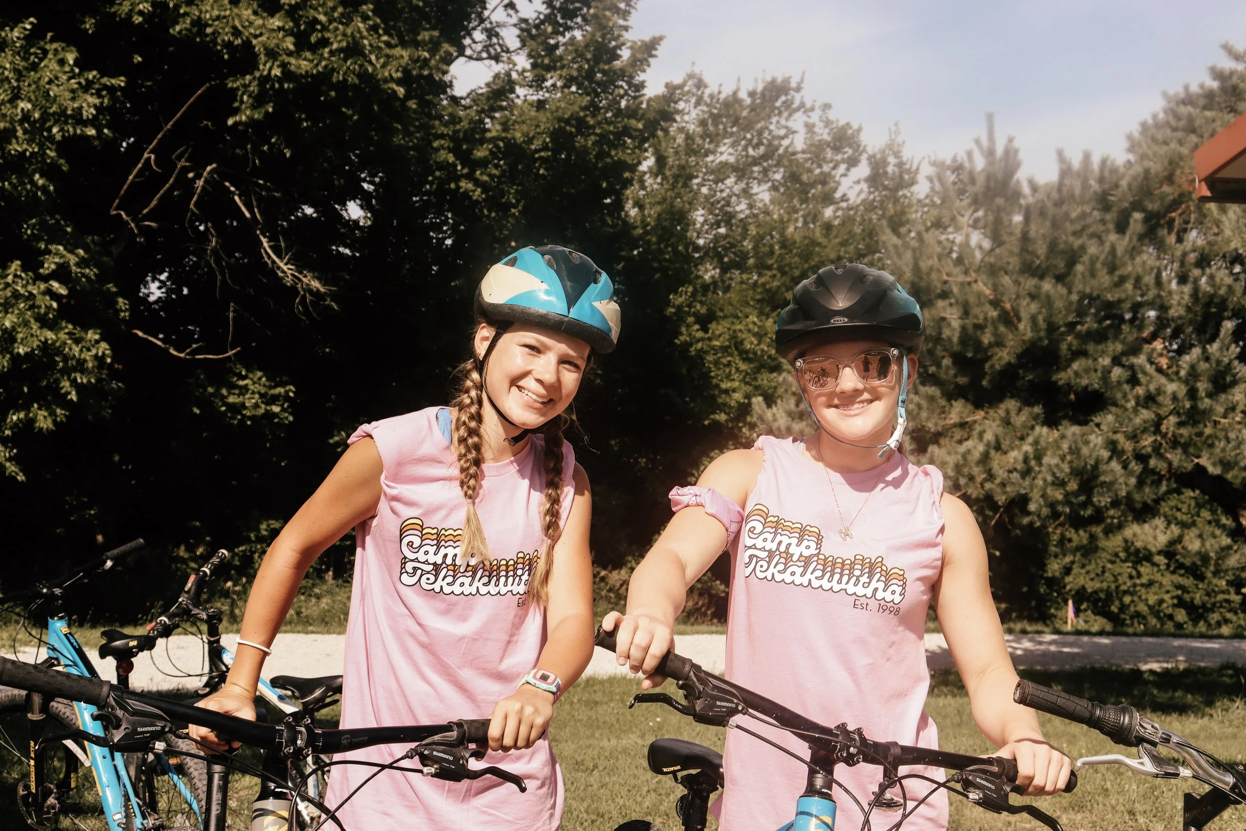 Two young women wearing helmets and pink T-shirts standing next to bicycles outdoors, smiling, with trees in the background.