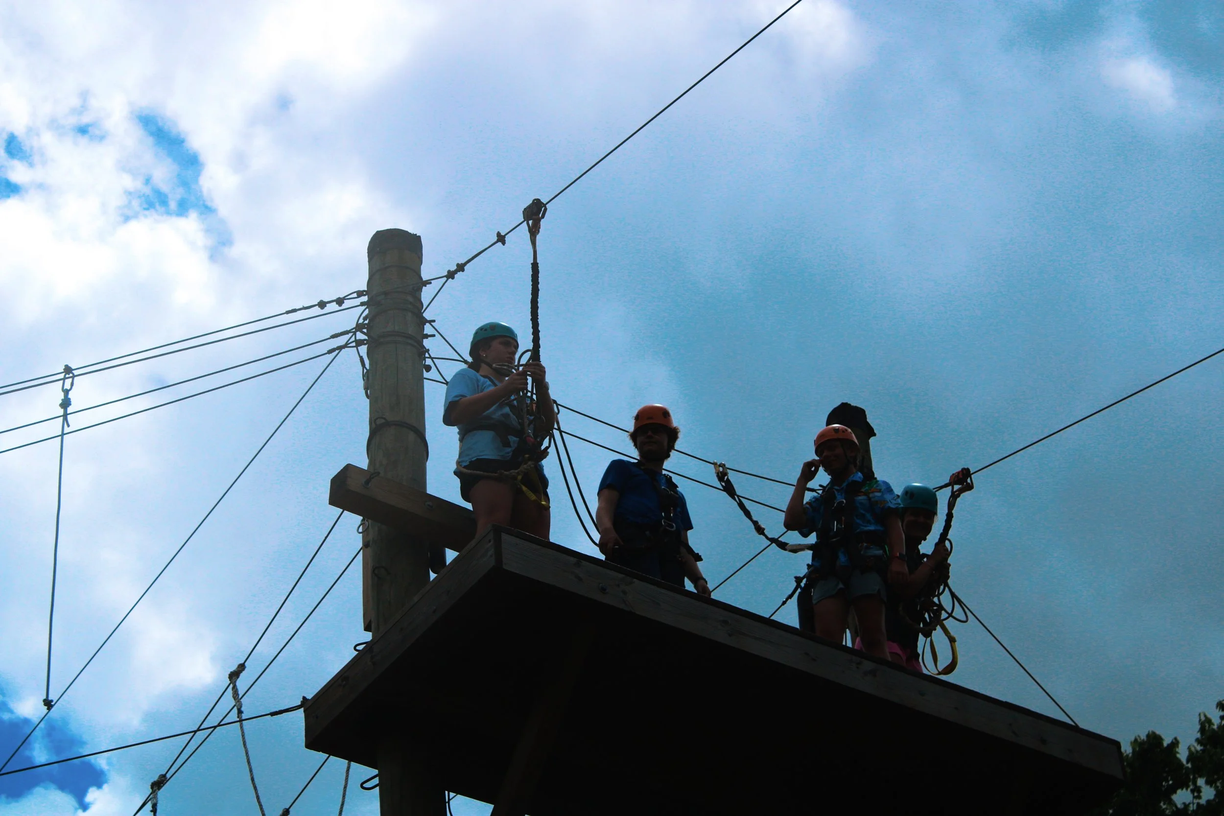 A group of people on a high ropes course wearing helmets and harnesses, standing on a wooden platform attached to a tall wooden pole, with blue sky and clouds in the background.