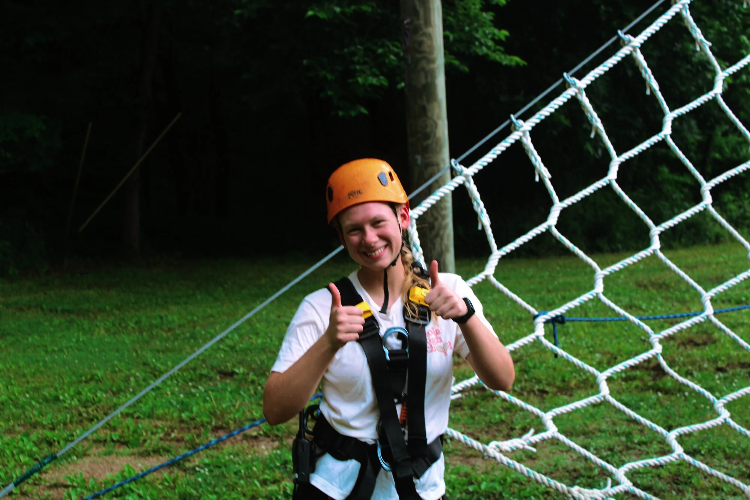 A young woman wearing a safety helmet and harness smiling and giving a thumbs-up in front of a climbing net in an outdoor adventure park.