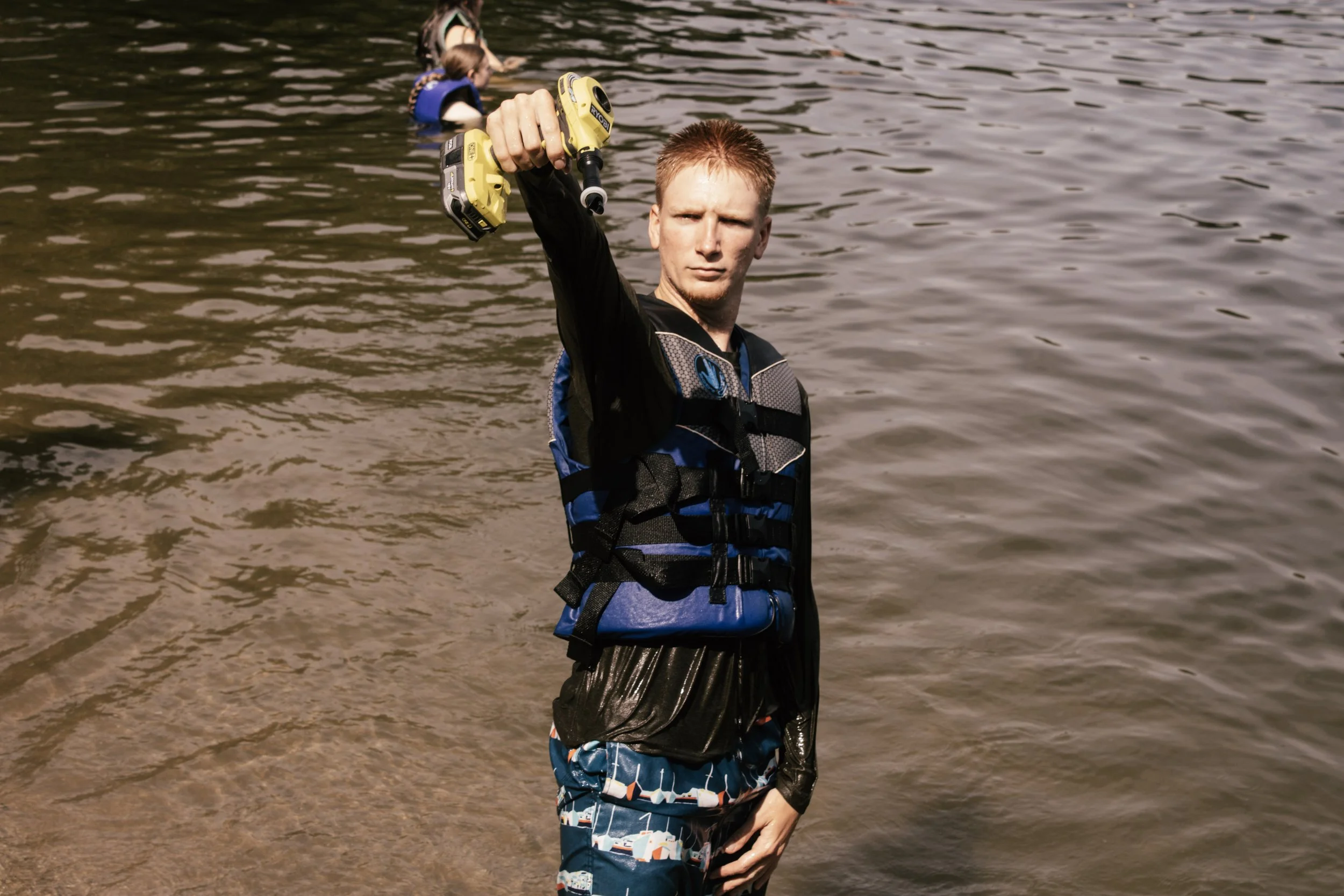 A young man stands in a body of water holding a yellow cordless drill in the air, with two children in the background swimming and playing in the water.