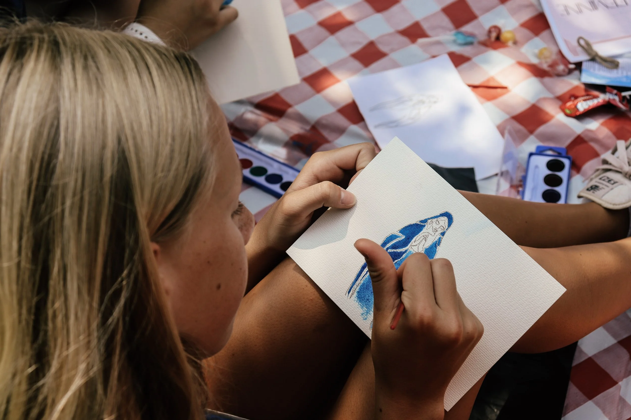A person with long blonde hair is painting a picture of a blue dolphin on a piece of white paper, while another person is nearby. The setting appears to be at a table covered with a red and white checkered tablecloth, with art supplies and drawings s