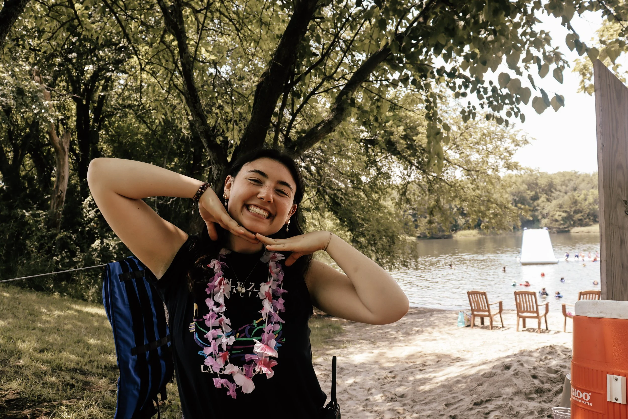 A young woman smiling with her hands under her chin, wearing a black shirt, pink lei, and beaded bracelet, outdoors near a lake with trees and people swimming in the background.