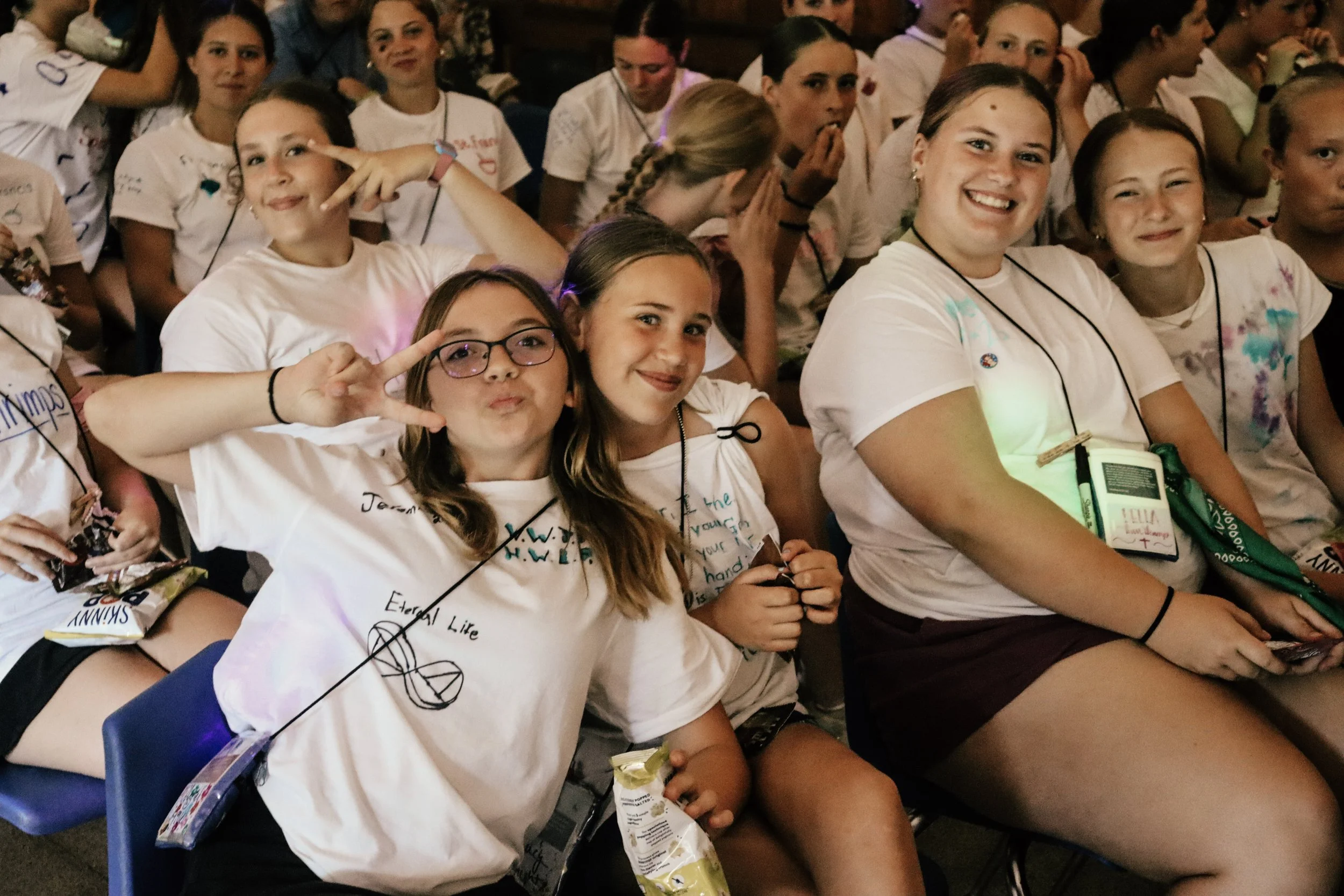 A group of smiling children sitting indoors, wearing casual white t-shirts, some making peace signs and others holding snacks and drinks, appearing happy and engaged in an event or gathering.