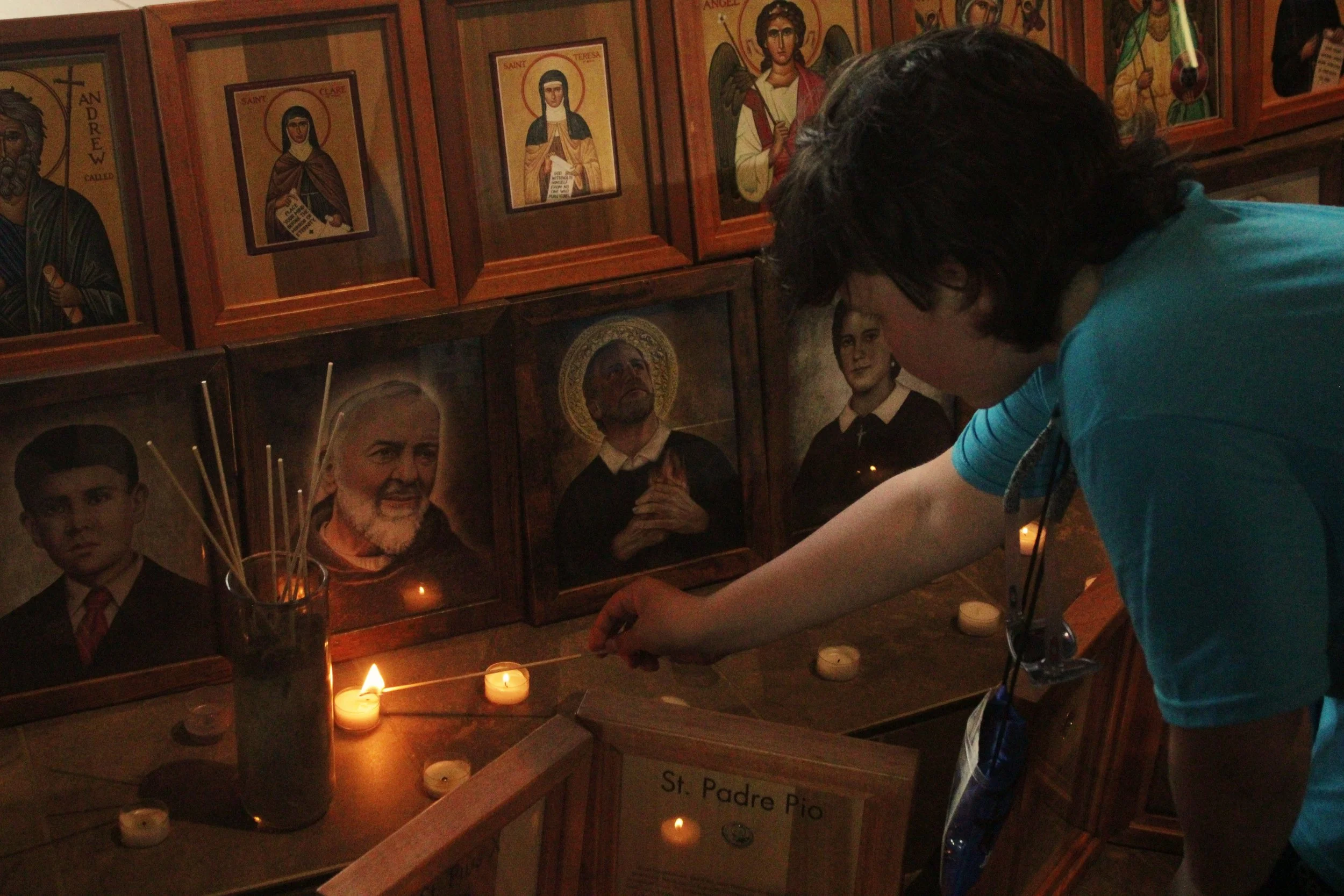 A person lighting candles in front of a wooden altar with framed religious icons and photographs of saints and religious figures. The altar has candles and incense sticks.