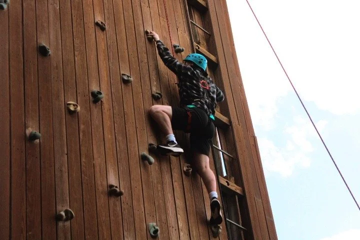 Child wearing a blue helmet climbing an indoor wooden rock wall with colorful handholds and footholds.