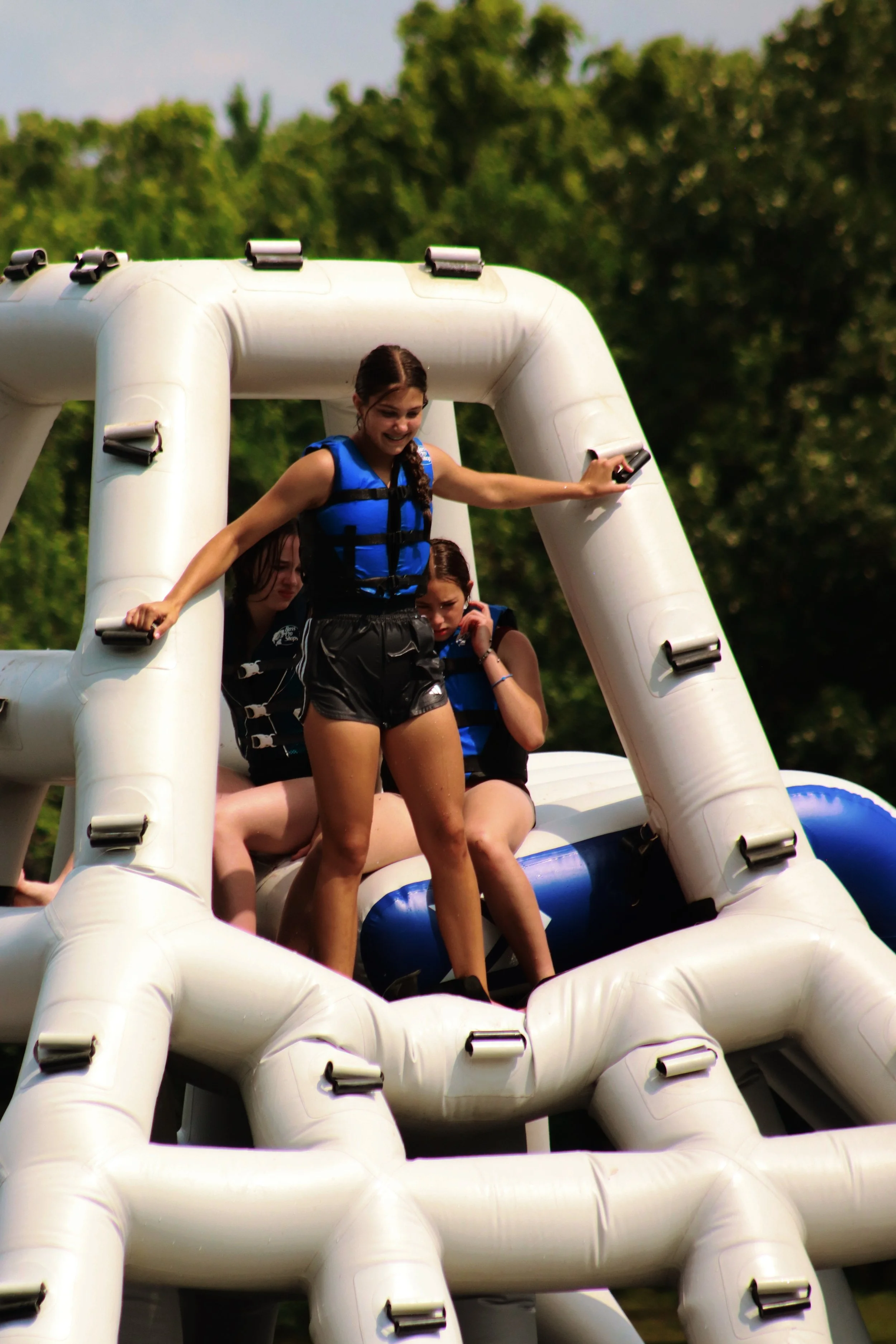 A group of people on a large inflatable water slide outdoors, with one girl in the front wearing a life vest, smiling and holding onto the slide, surrounded by greenery and trees.