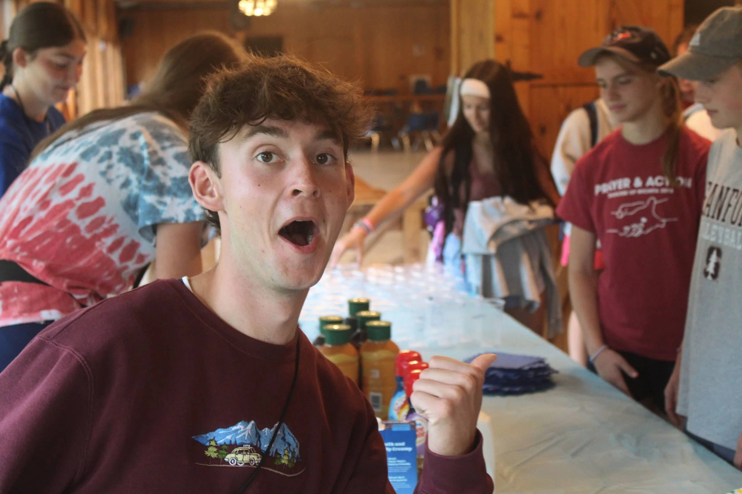 A young man with dark hair and a surprised expression on his face, wearing a maroon sweatshirt with a graphic, looking at the camera. He is indoors at a table with several bottles of sauces or condiments, and other people in the background are engagi